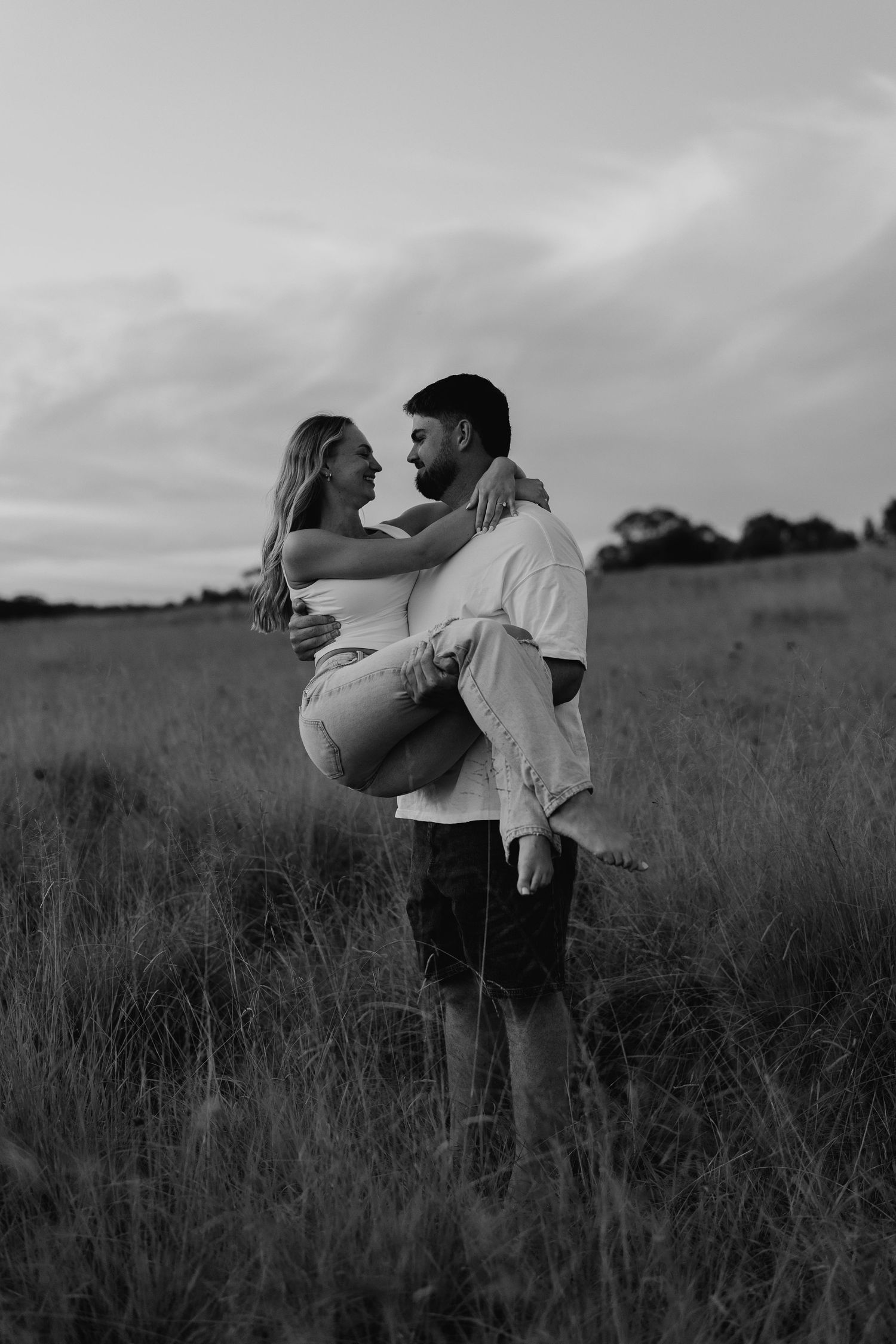 Black and white photo of man lifting woman in his arms in grassy field at dusk, both smiling at each other.