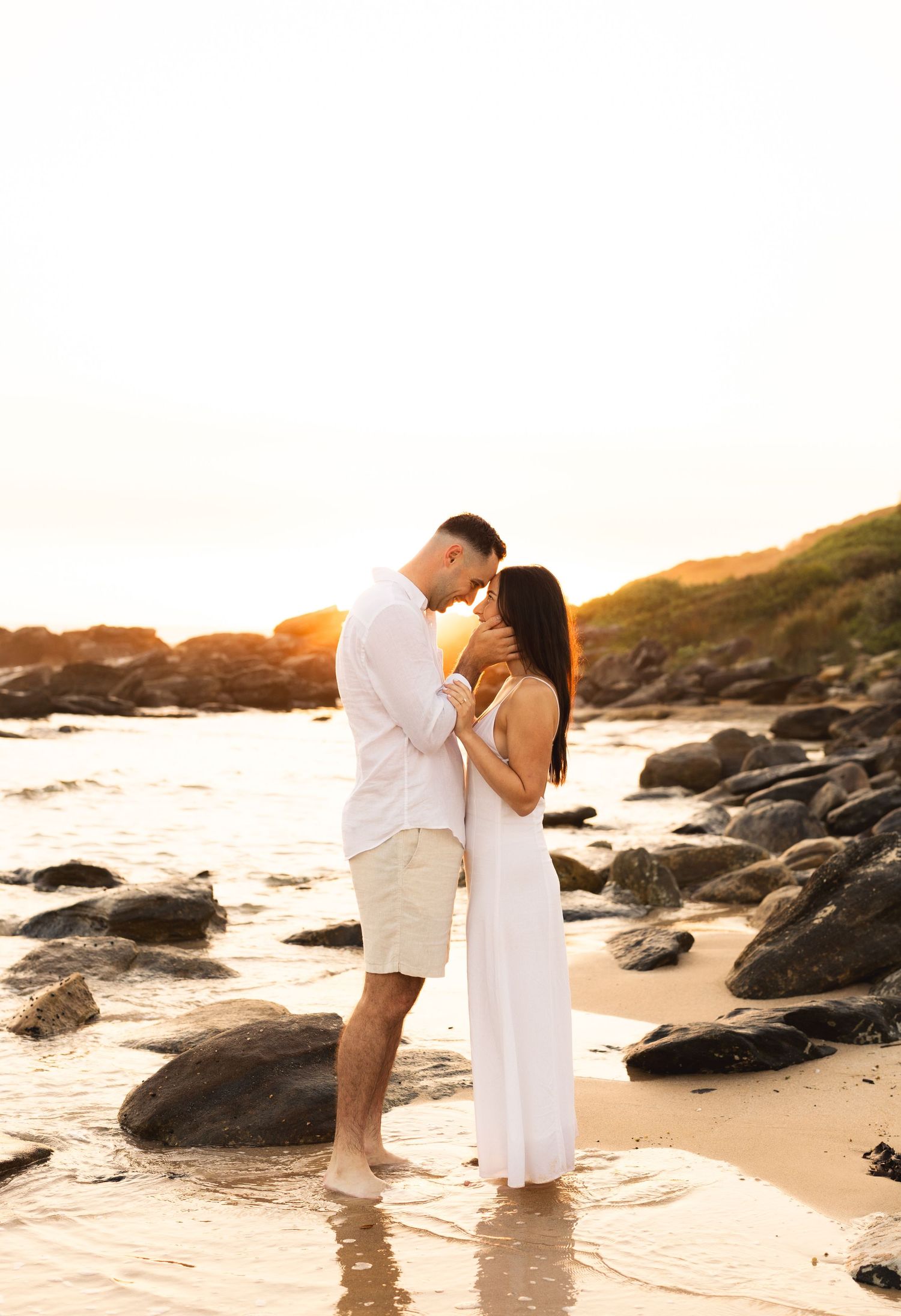 Romantic couple standing close together on a rocky beach shore during a warm golden sunset.