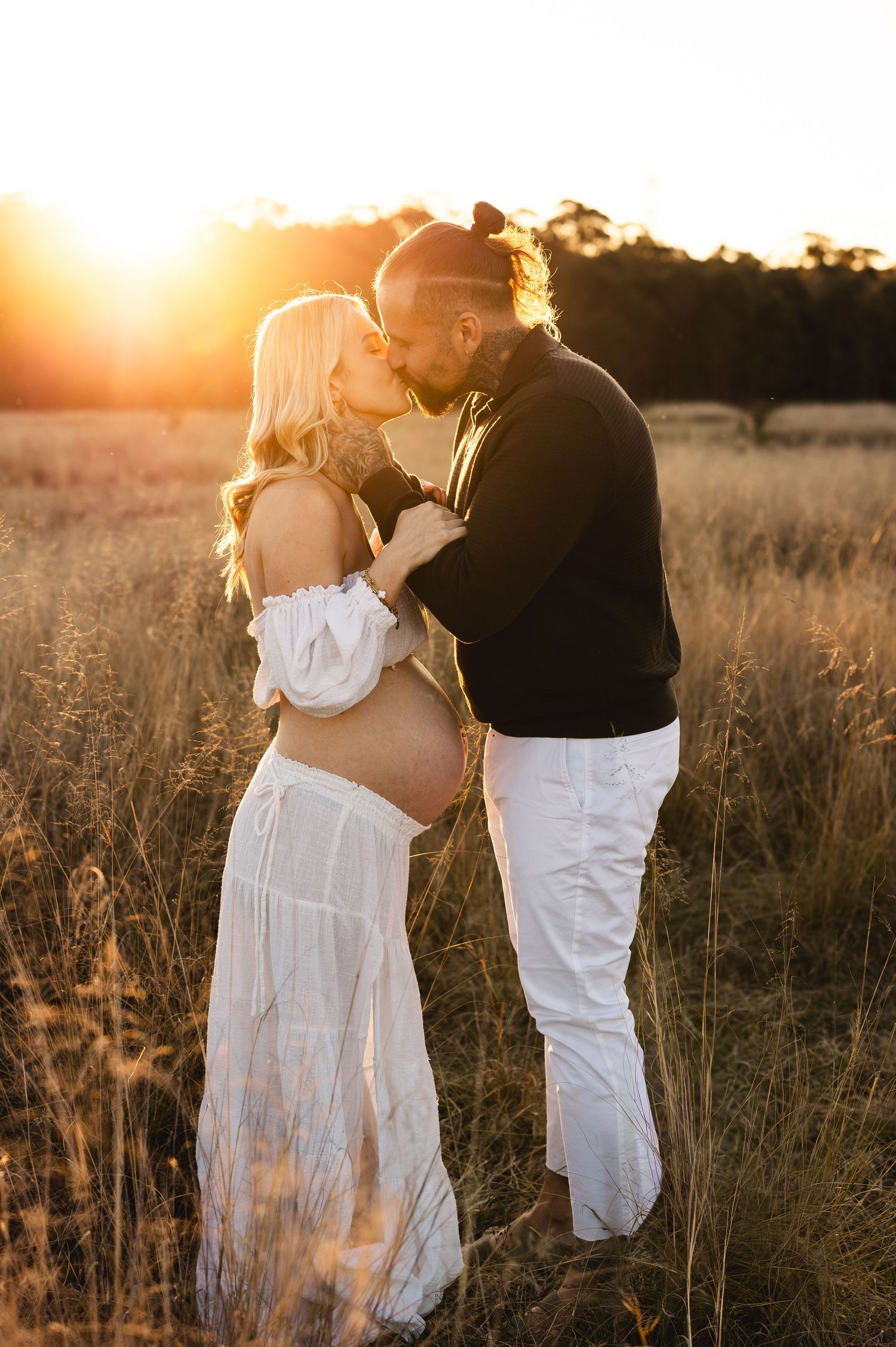 Pregnant couple sharing a kiss in golden field at sunset, woman's baby bump visible.