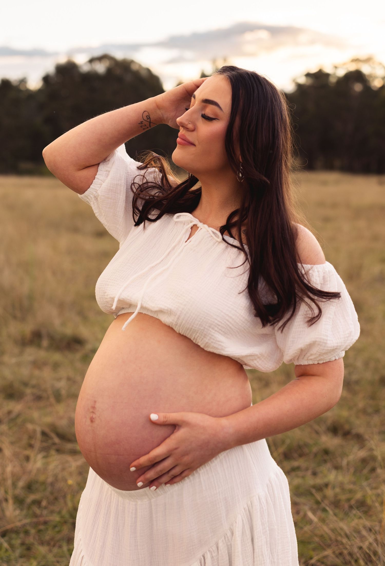 Pregnant woman in a white dress cradling her baby bump at golden hour in an open field.