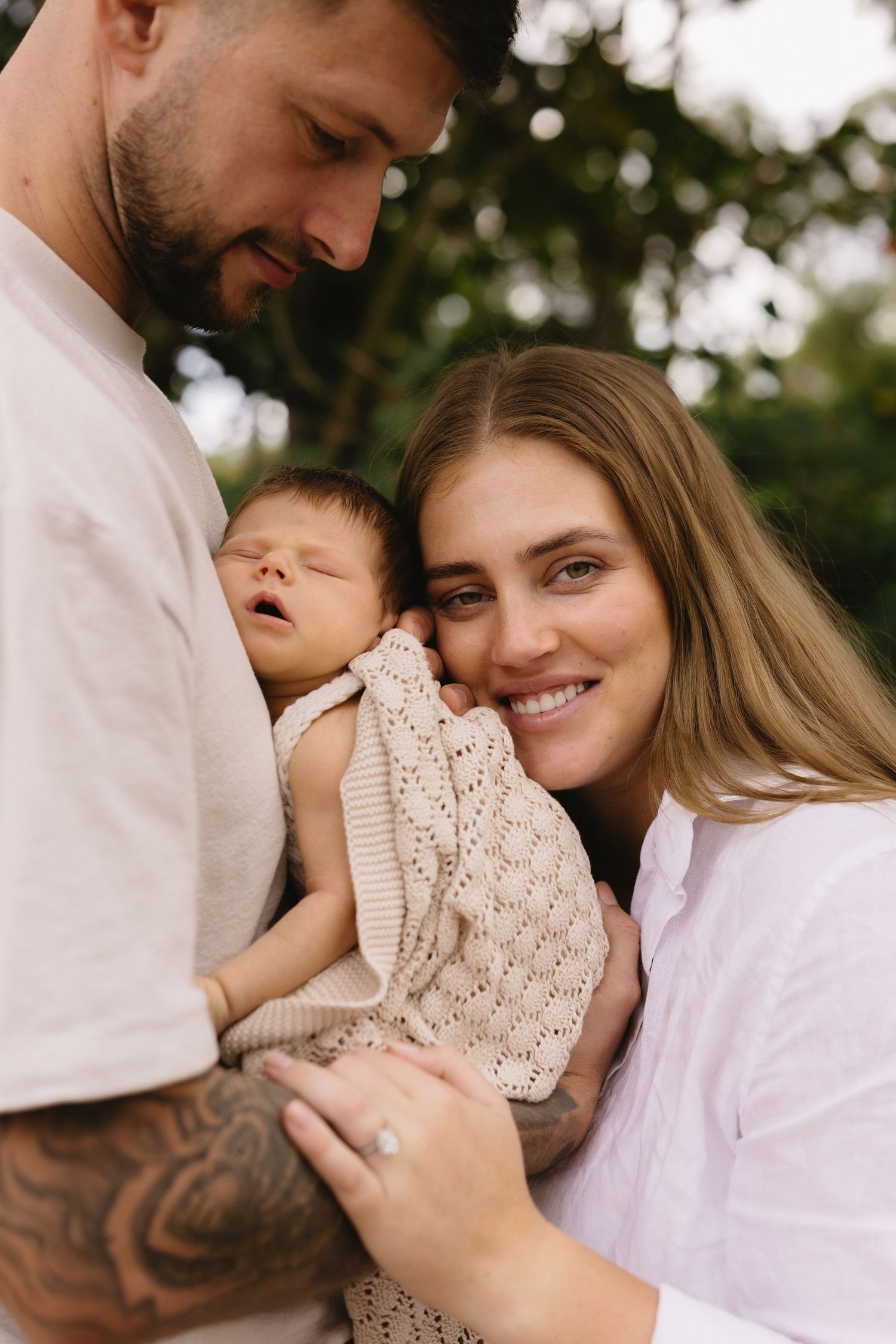 A couple smiles while cradling their newborn baby outdoors, with the father's tattooed arm visible.