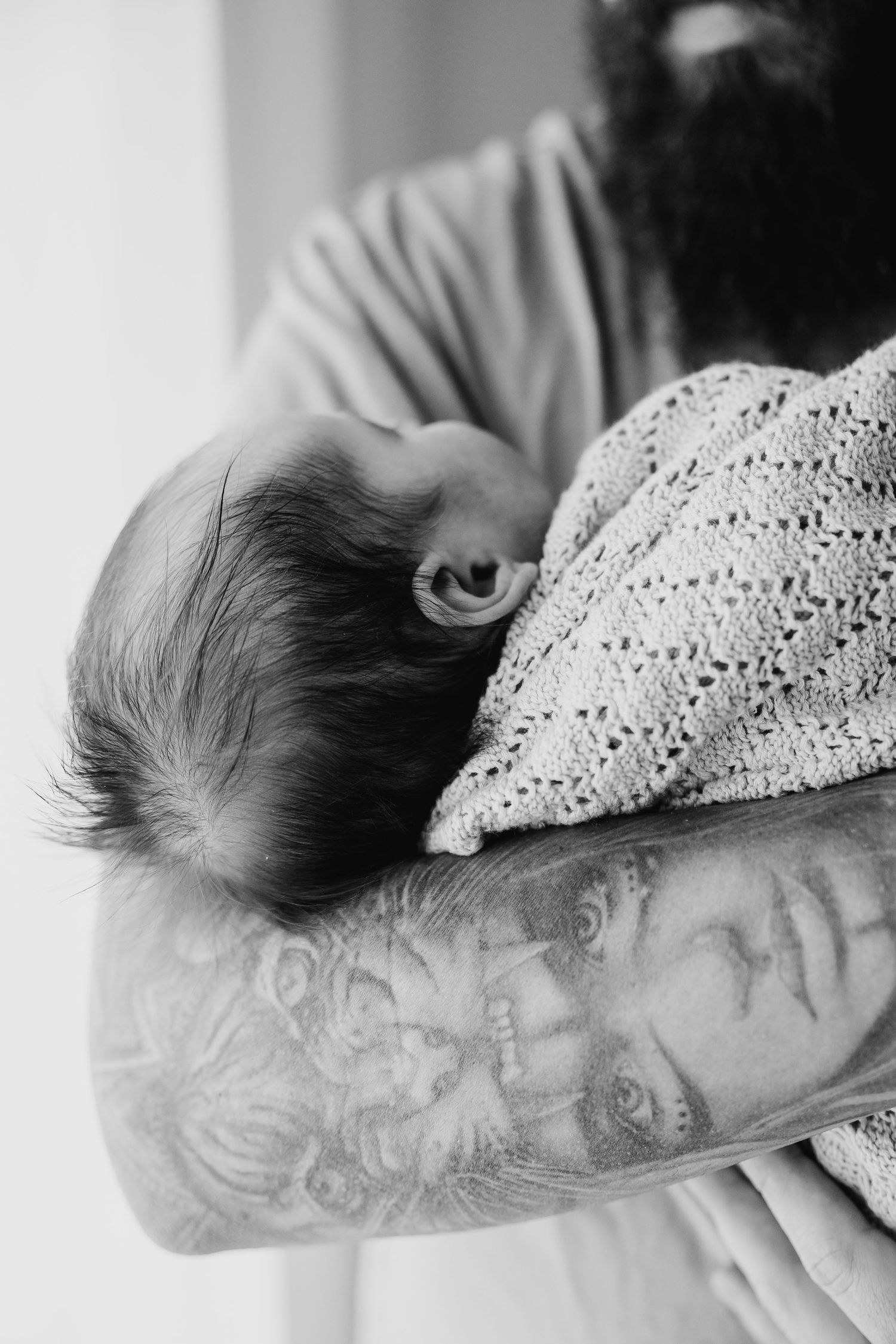 Close-up black and white image of a tattooed father holding a sleeping newborn baby.