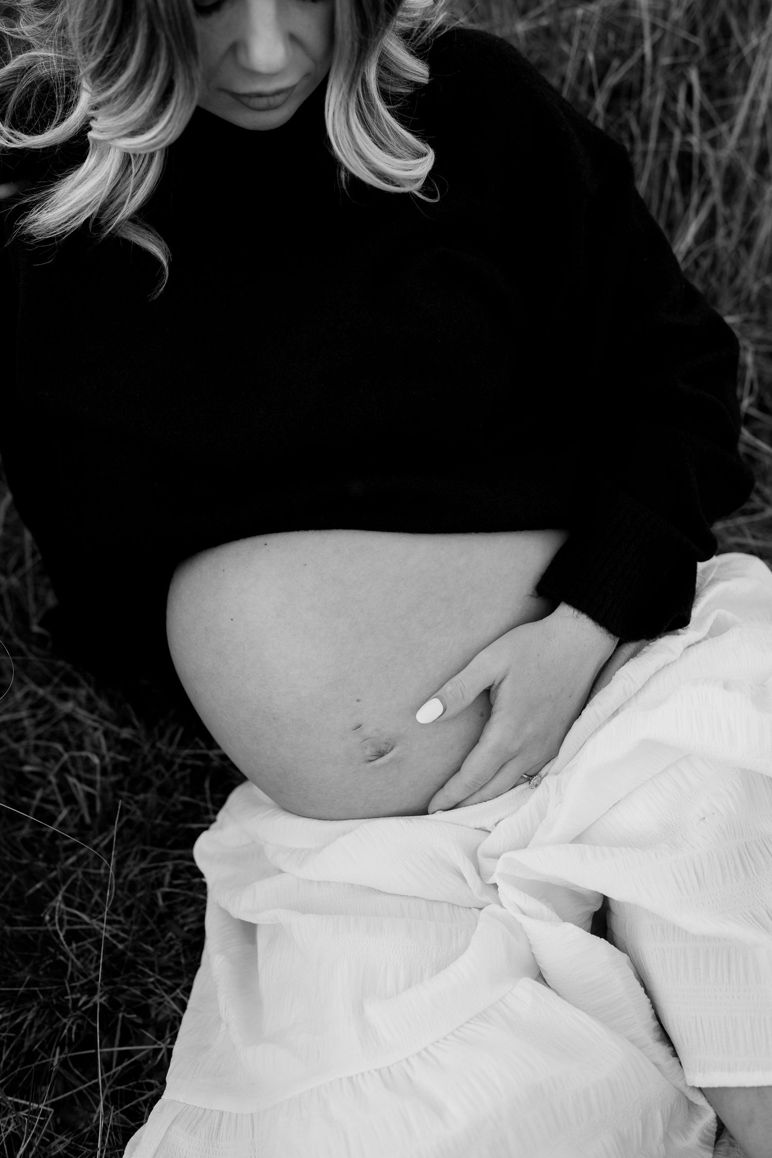 Black and white close-up of pregnant woman sitting in field, gently holding her bare baby bump.