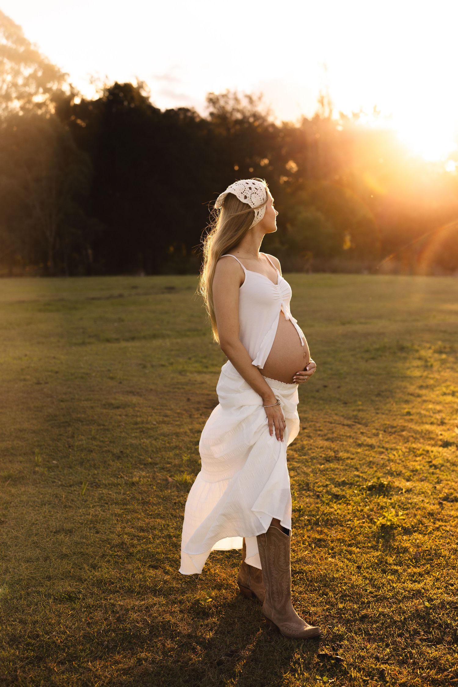 Pregnant woman in white dress and headband at golden hour sunset in park.