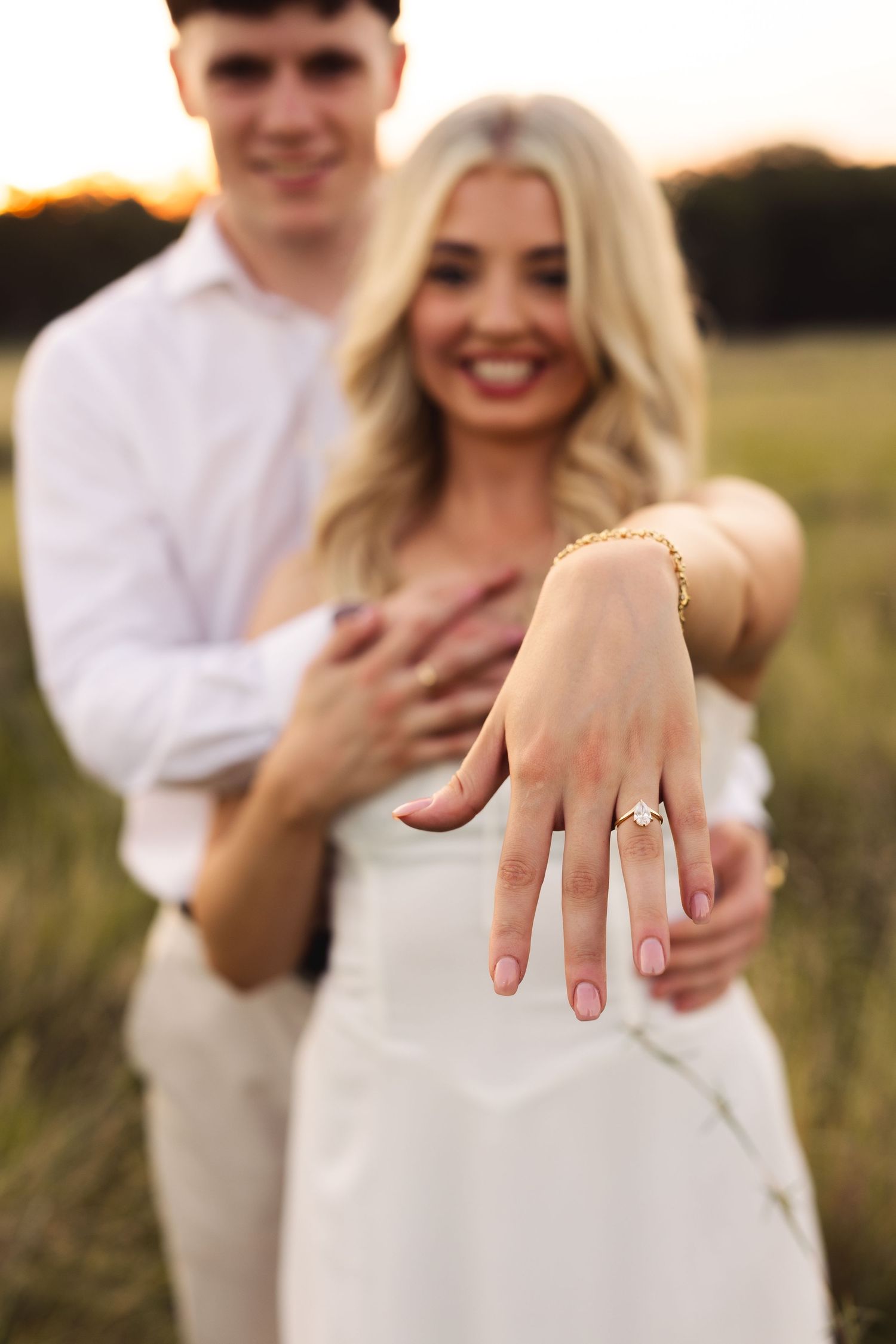Smiling blonde woman shows off engagement ring while partner embraces her from behind in a field.