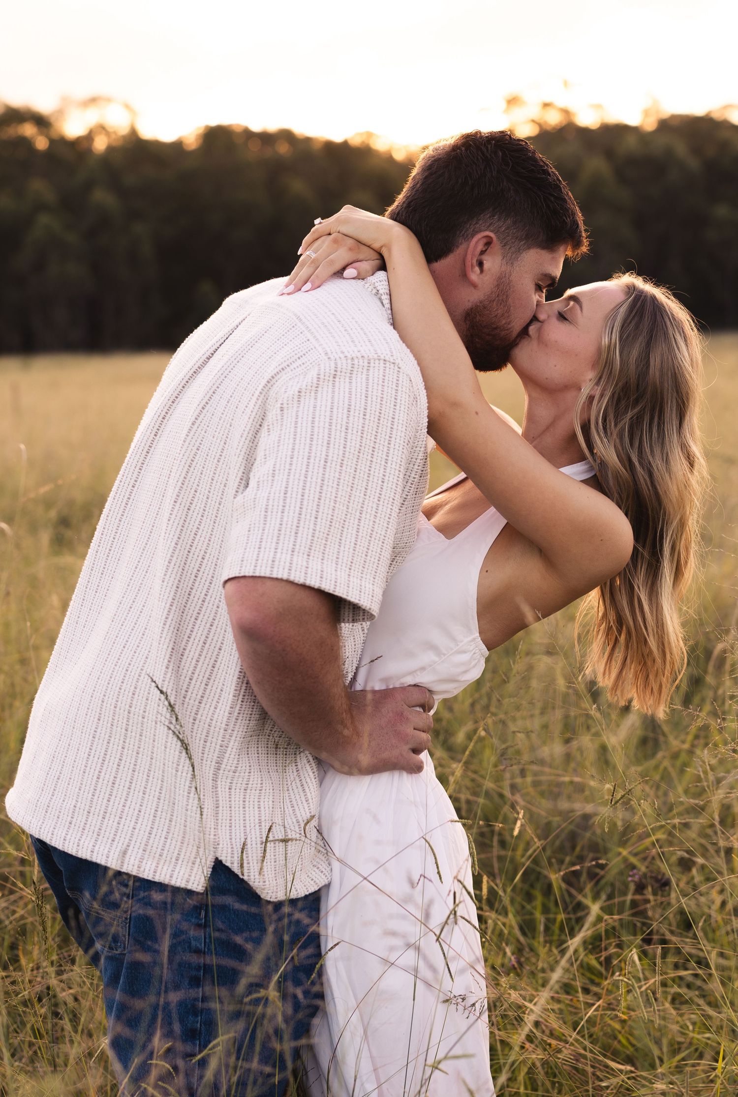 Couple sharing a passionate kiss at golden hour in tall grass field, woman in white dress, man in striped shirt.