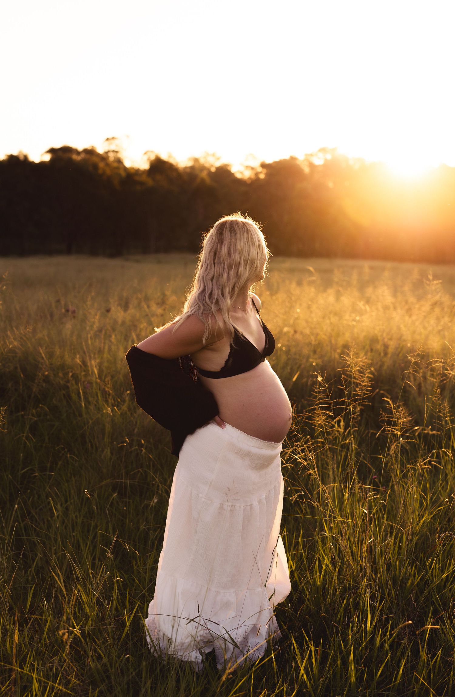 Pregnant blonde woman in white skirt and black top standing in sunlit field.