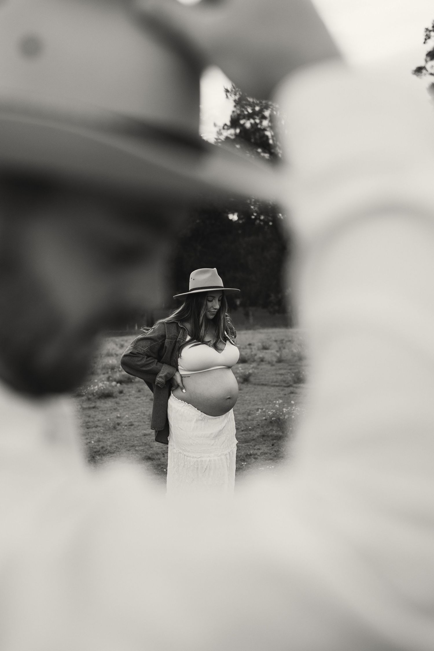 Black and white maternity photo of pregnant woman in hat and white dress in rural setting.