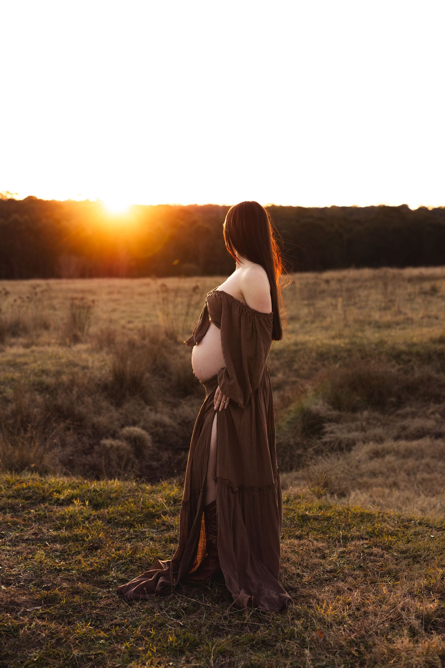 Pregnant woman in brown off-shoulder dress standing in golden sunset field, cradling baby bump during maternity photoshoot.