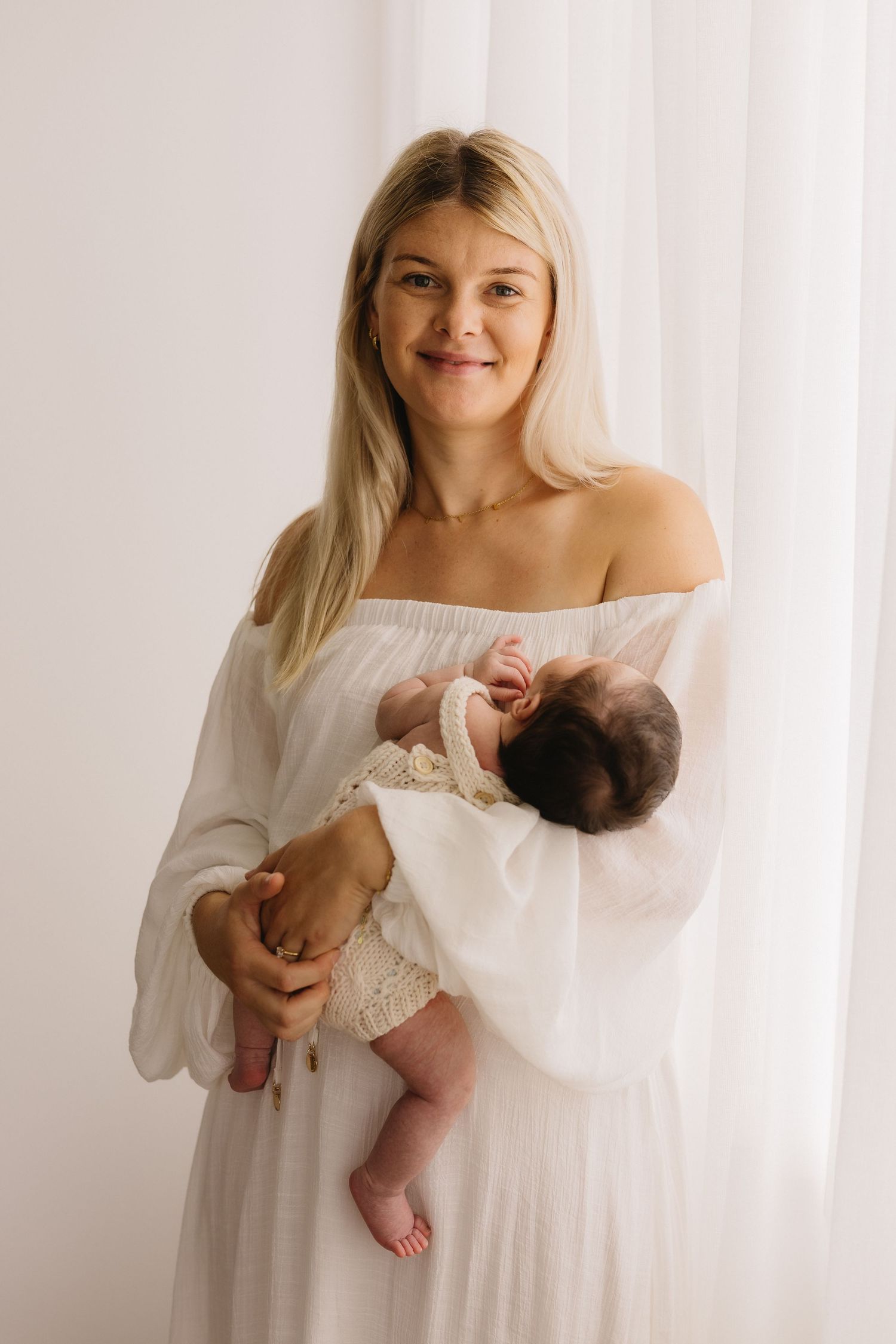 Smiling blonde mother in white off-shoulder dress holding dark-haired newborn baby by window.