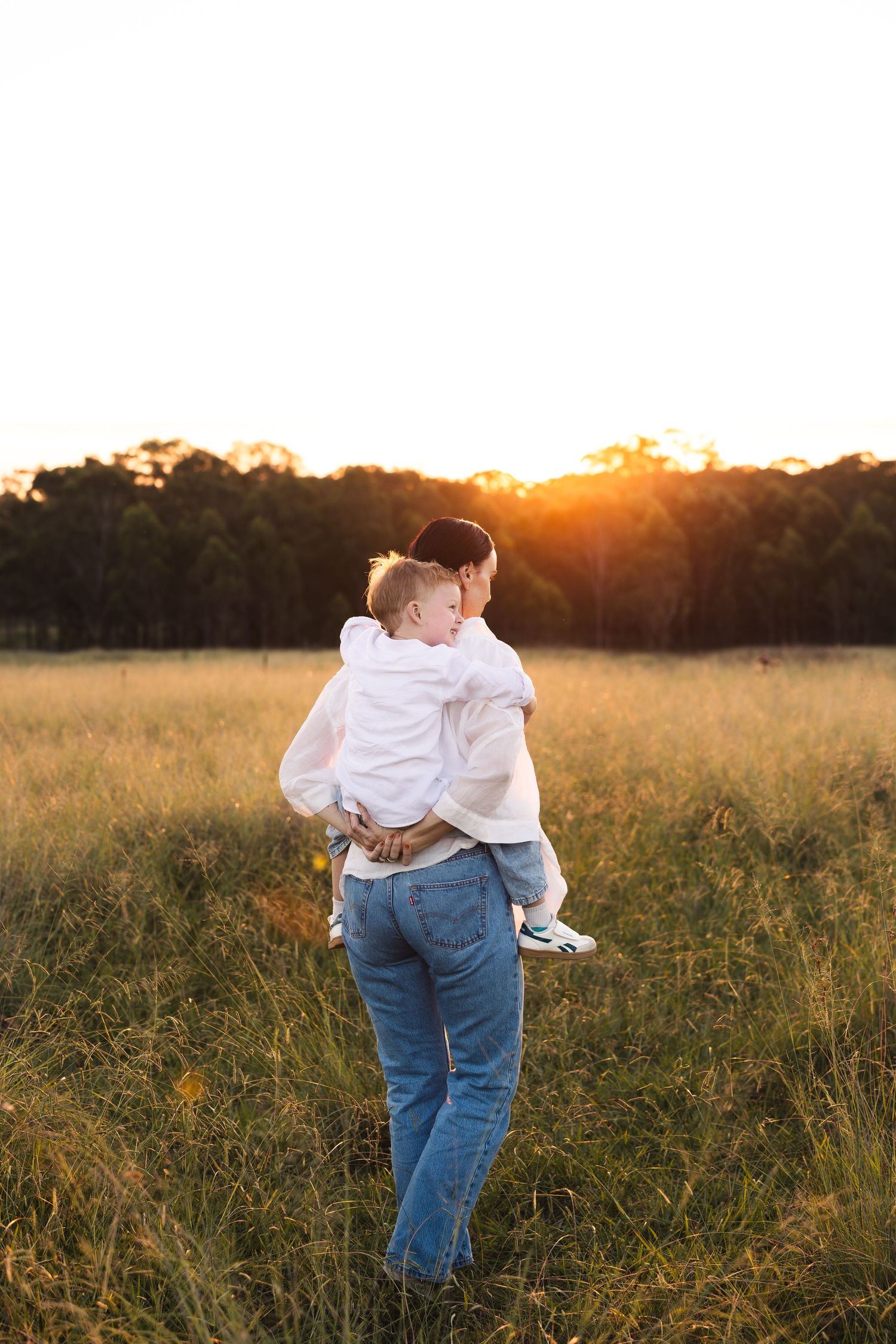 Mother holding young child while standing in golden sunlit field at sunset.
