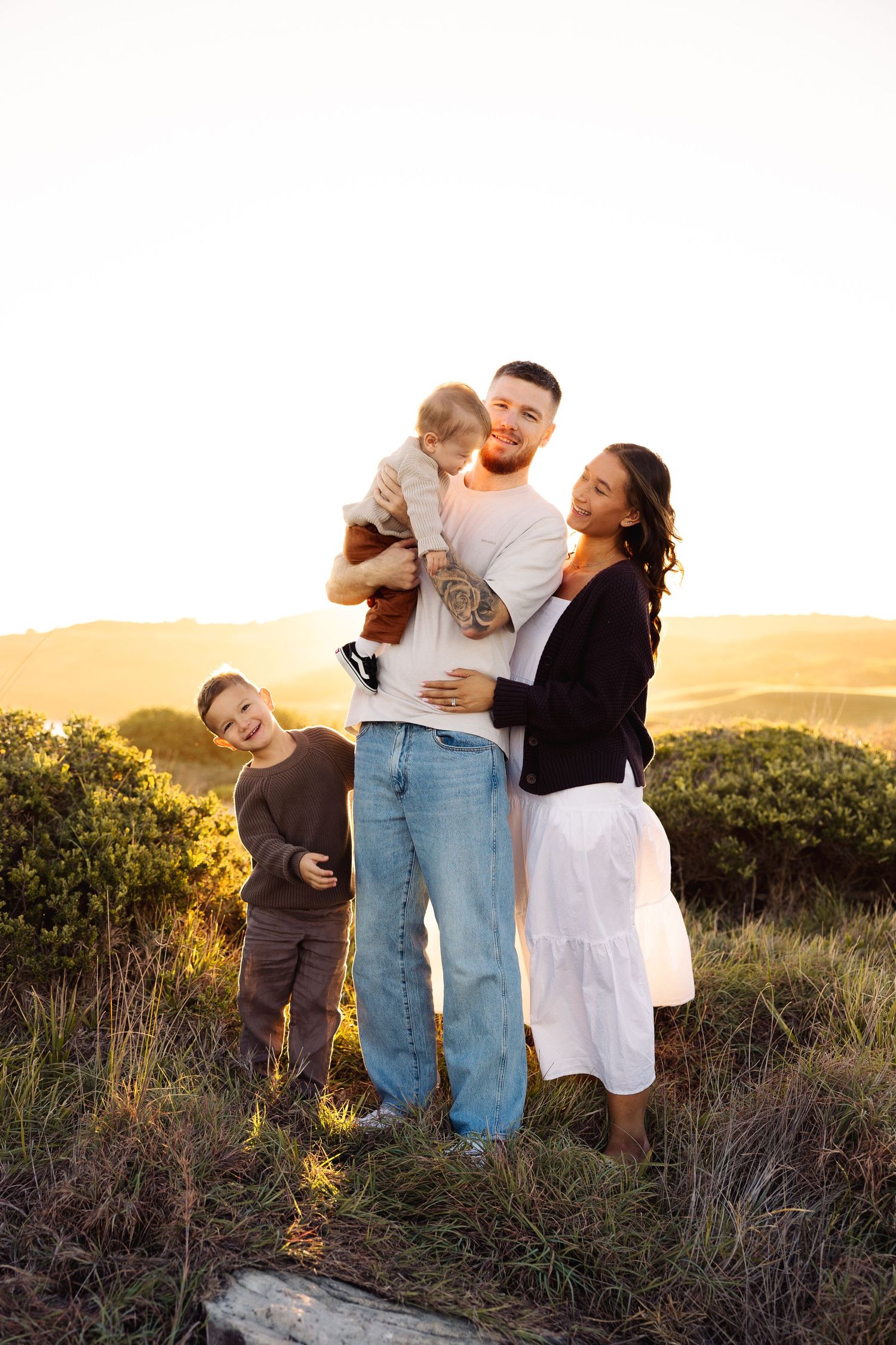 Family of four posing at golden sunset on a hill, father holding baby, son beside them.