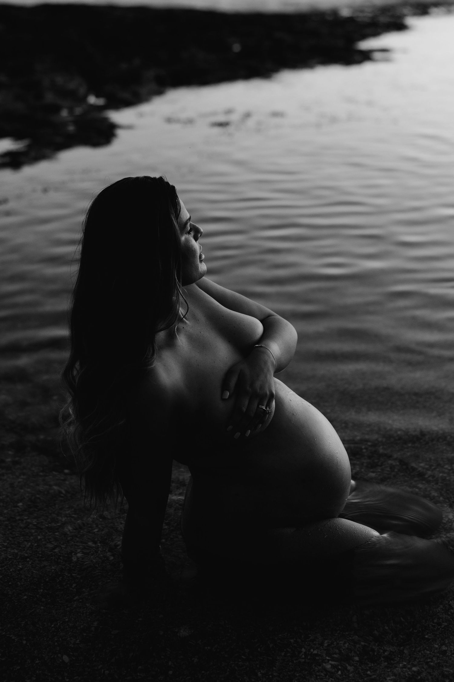 Black and white artistic photo of pregnant woman sitting by water at sunset.