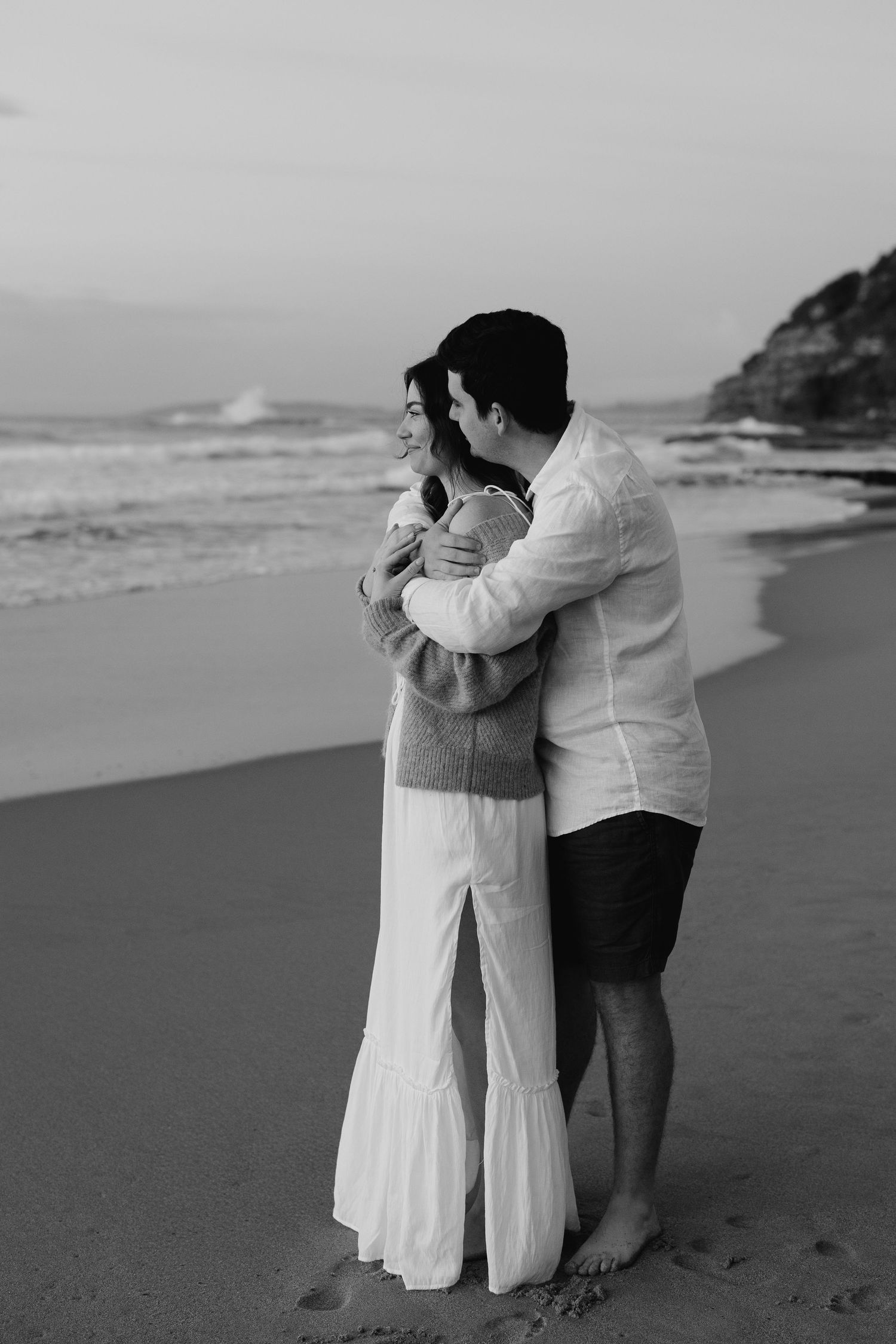 Black and white romantic couple embracing on beach at sunset, woman in white flowy dress, man in white shirt.