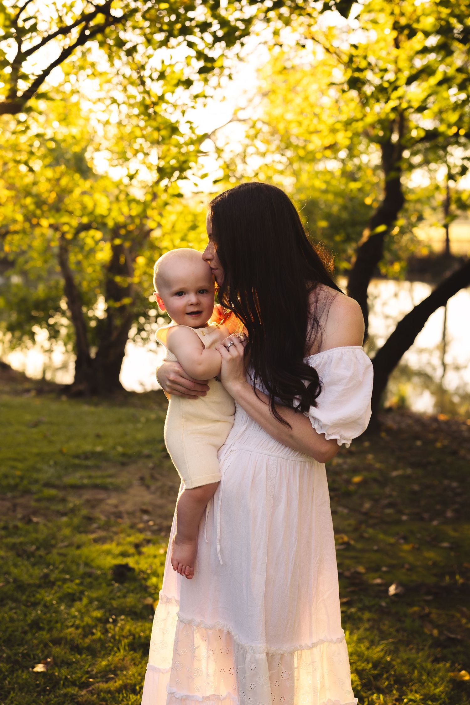 A mother in a white dress kisses her baby outdoors in golden sunlight surrounded by lush green trees.