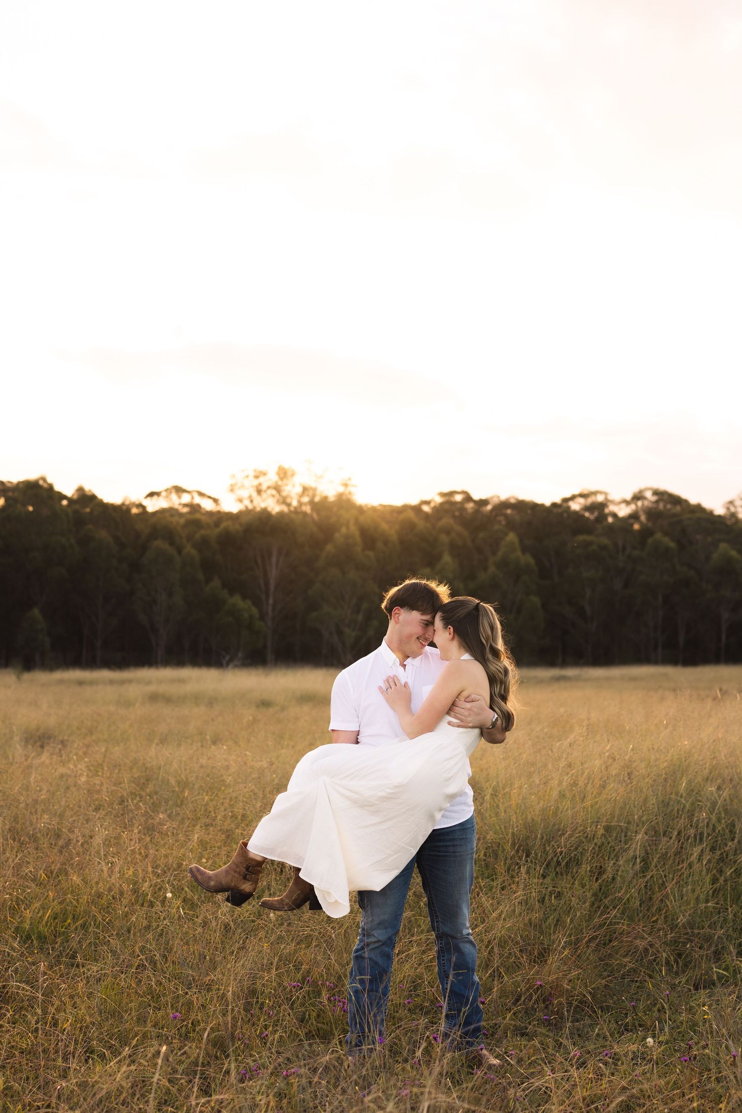 A man carries a woman in a white dress through a golden field at sunset, sharing a romantic moment.