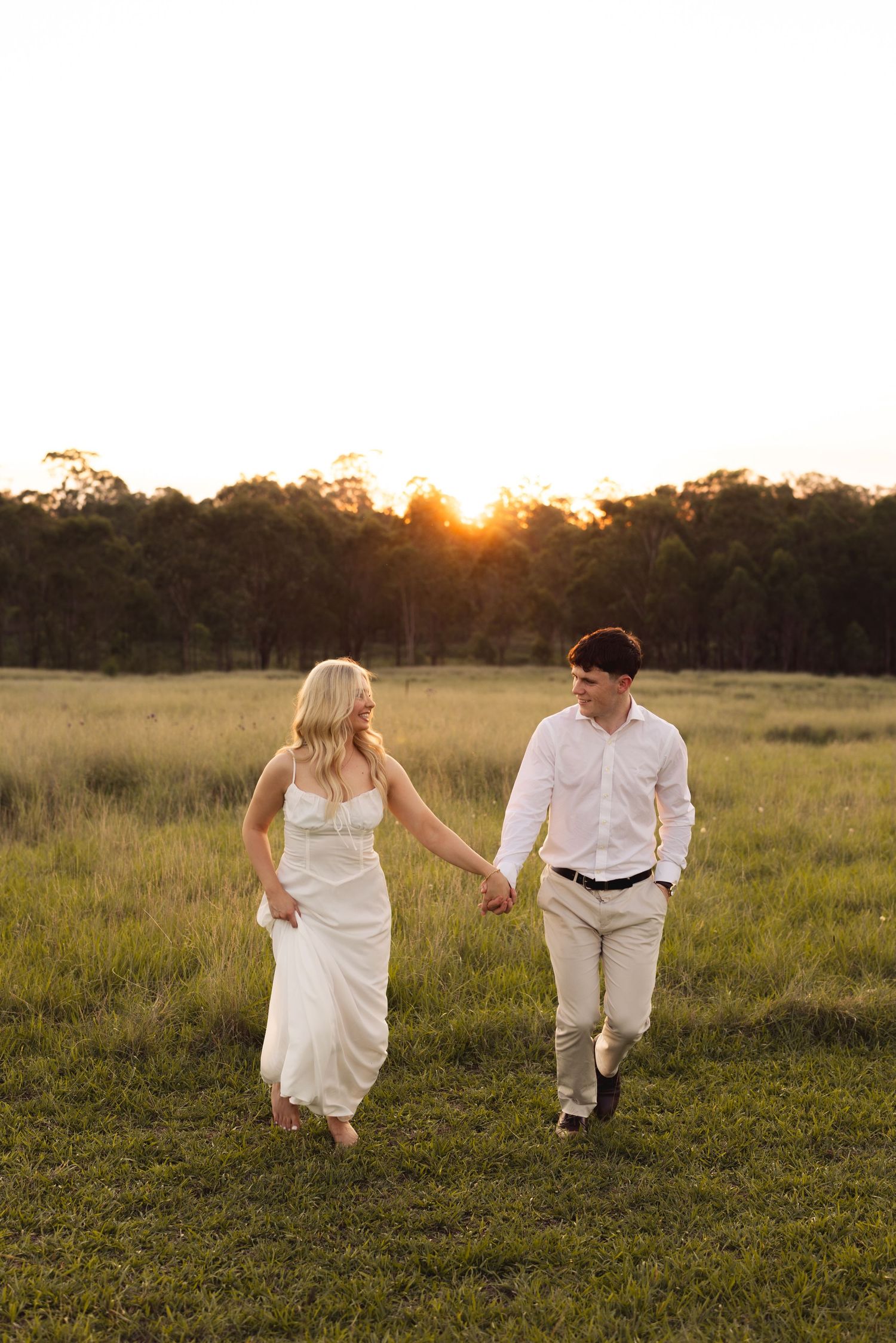 Joyful couple holding hands and walking through a sunlit green field at golden hour sunset.