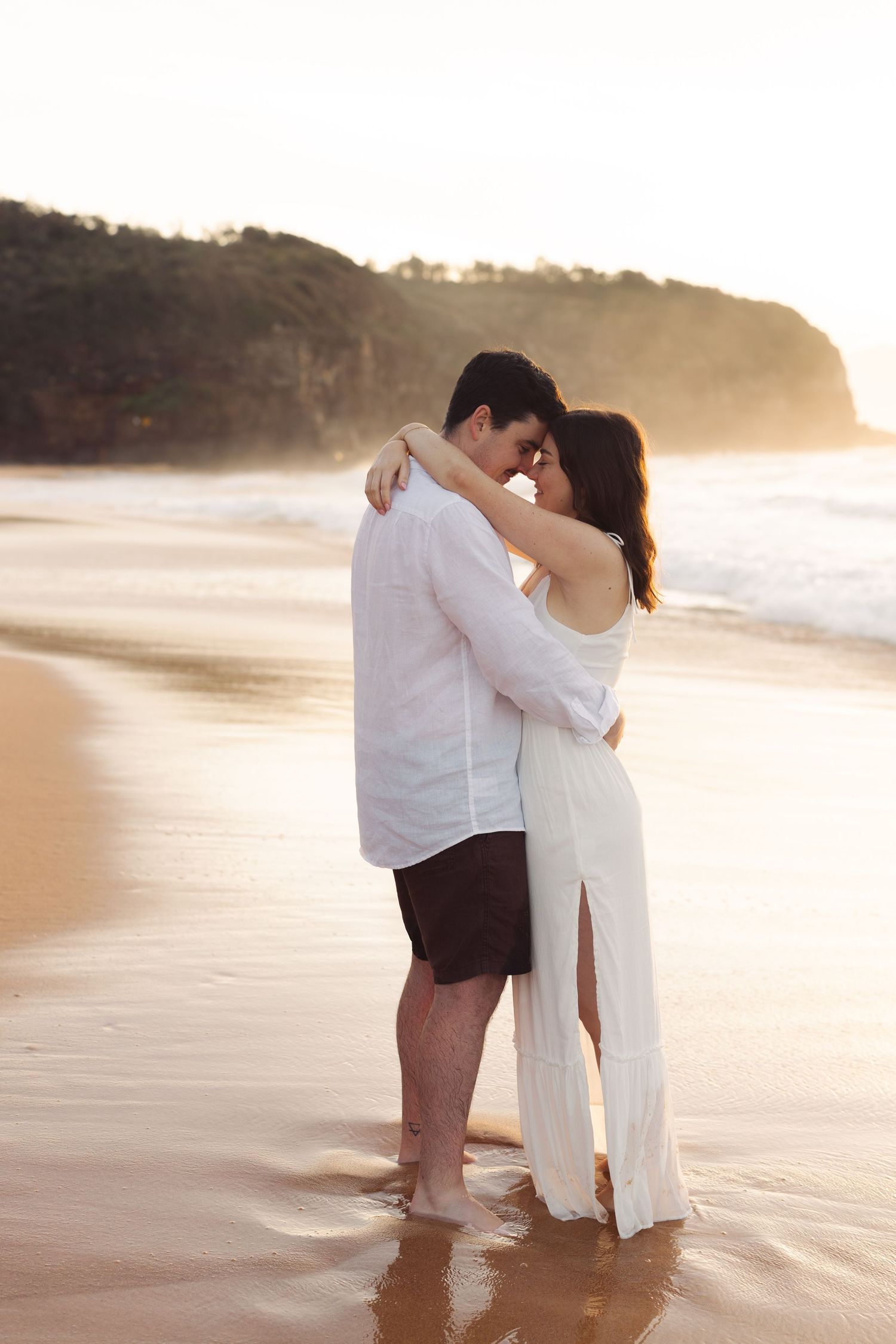 Couple embracing barefoot on golden sandy beach at sunset, woman in white slit dress, man in white shirt.