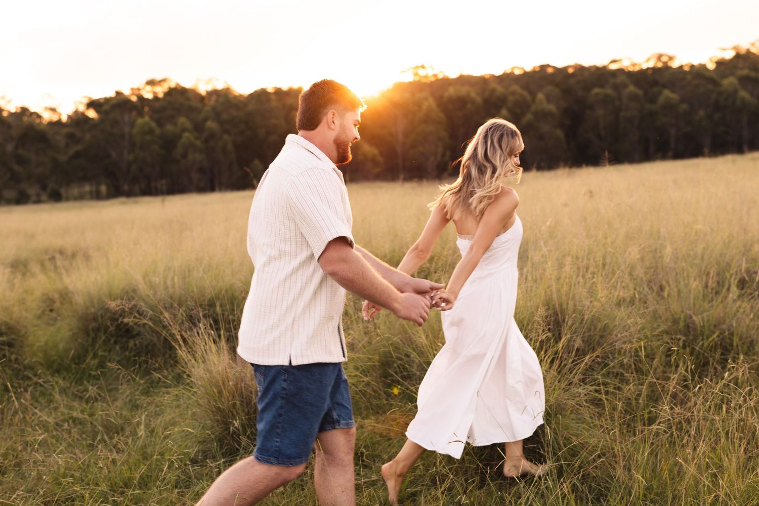 Couple holding hands walking through golden sunlit meadow at sunset, woman in white dress, man in blue shorts.