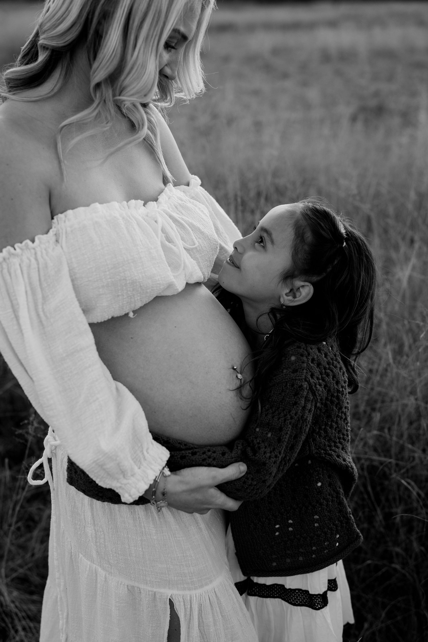 Black and white photo of young girl hugging pregnant mother's baby bump in a field.