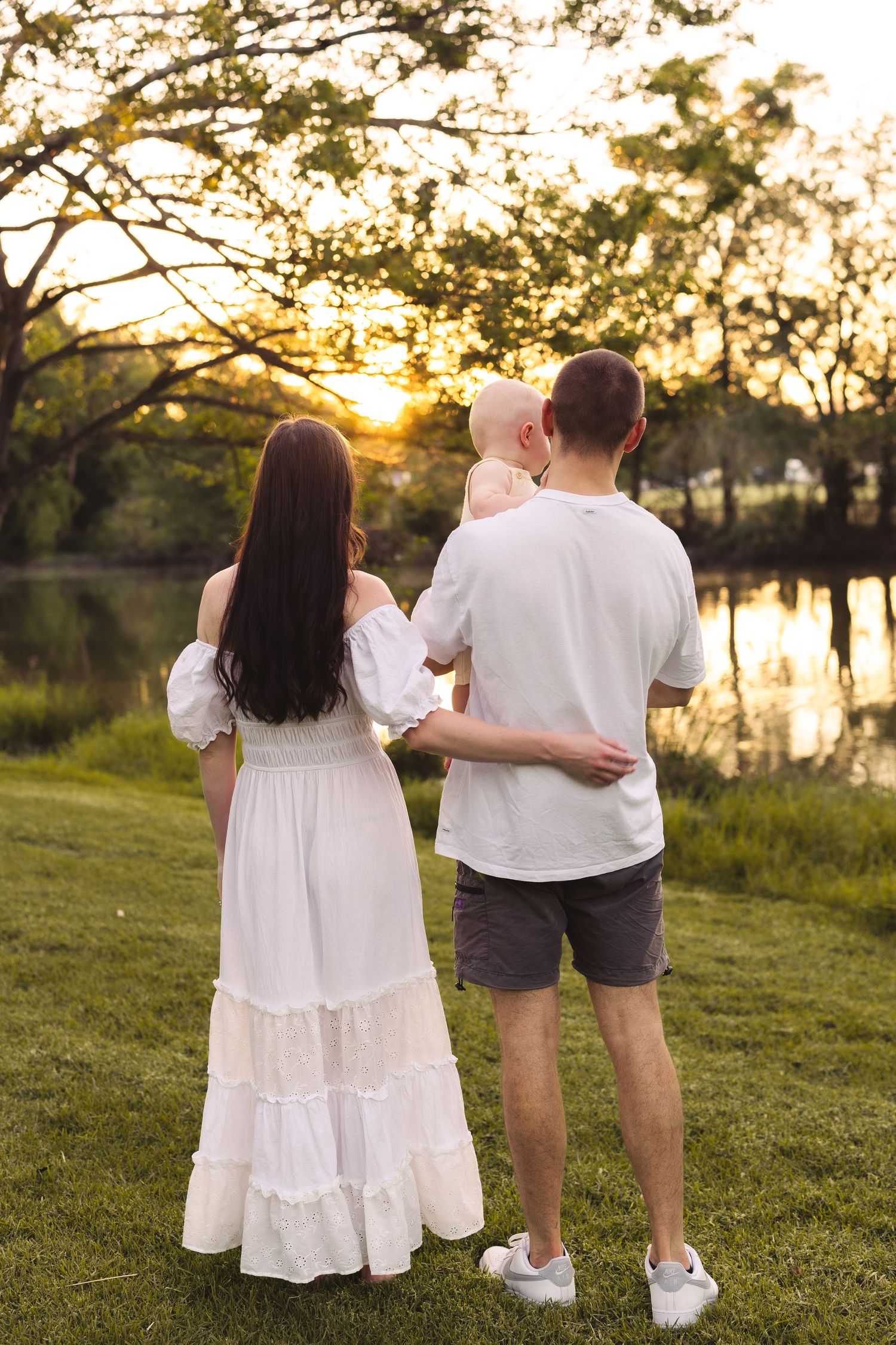 Young family with baby enjoying sunset by a lake in a park, mother in white dress, father in white shirt.