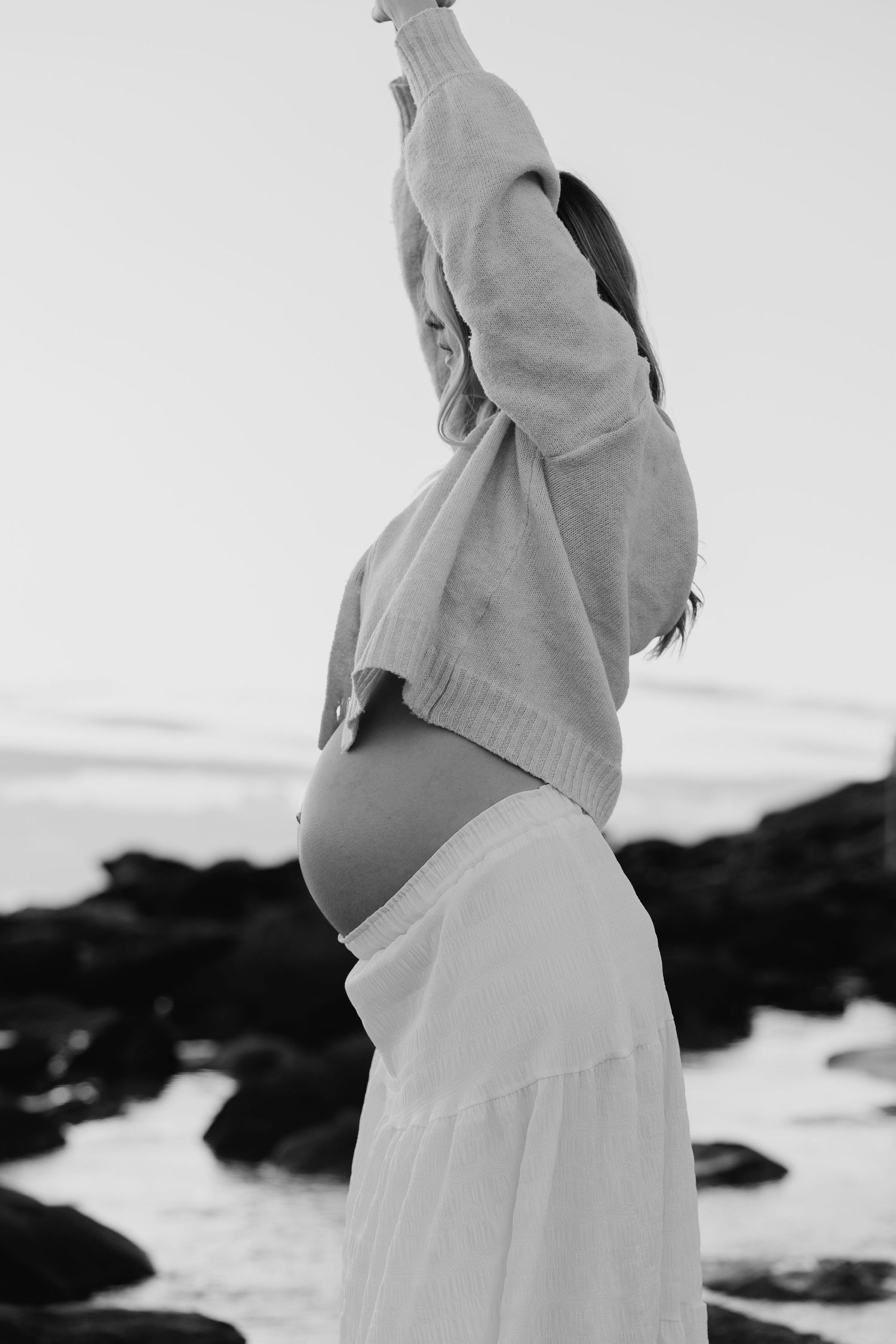 Black and white photo of pregnant woman at beach stretching arms up, showing bare baby bump between sweater and skirt.