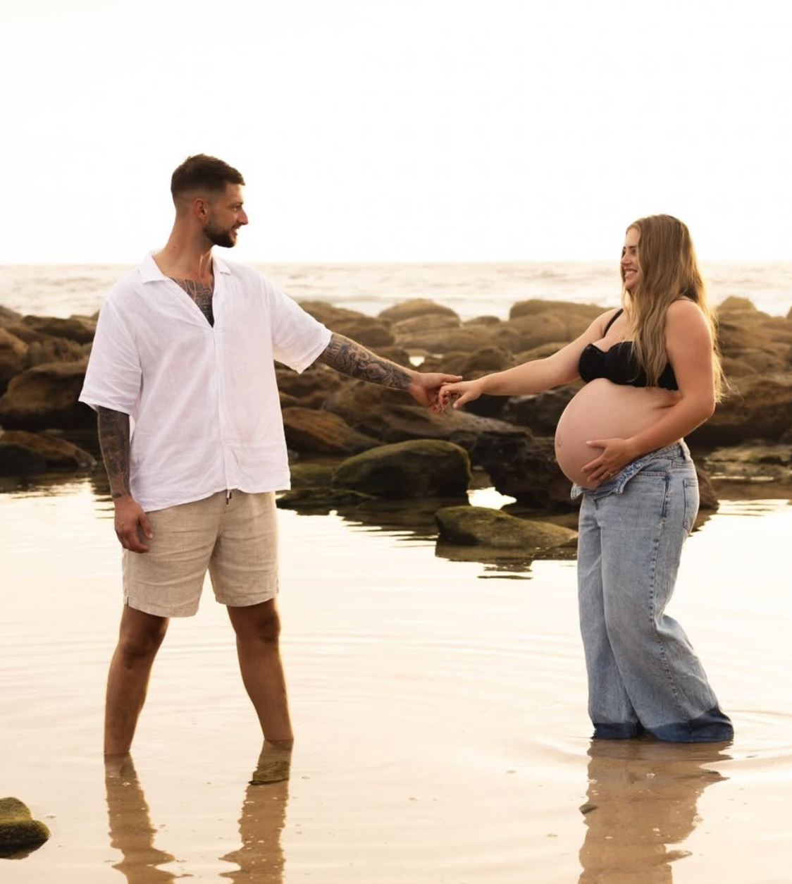 Couple holding hands on beach, pregnant woman in black top and jeans showing baby bump, man in white shirt and shorts.