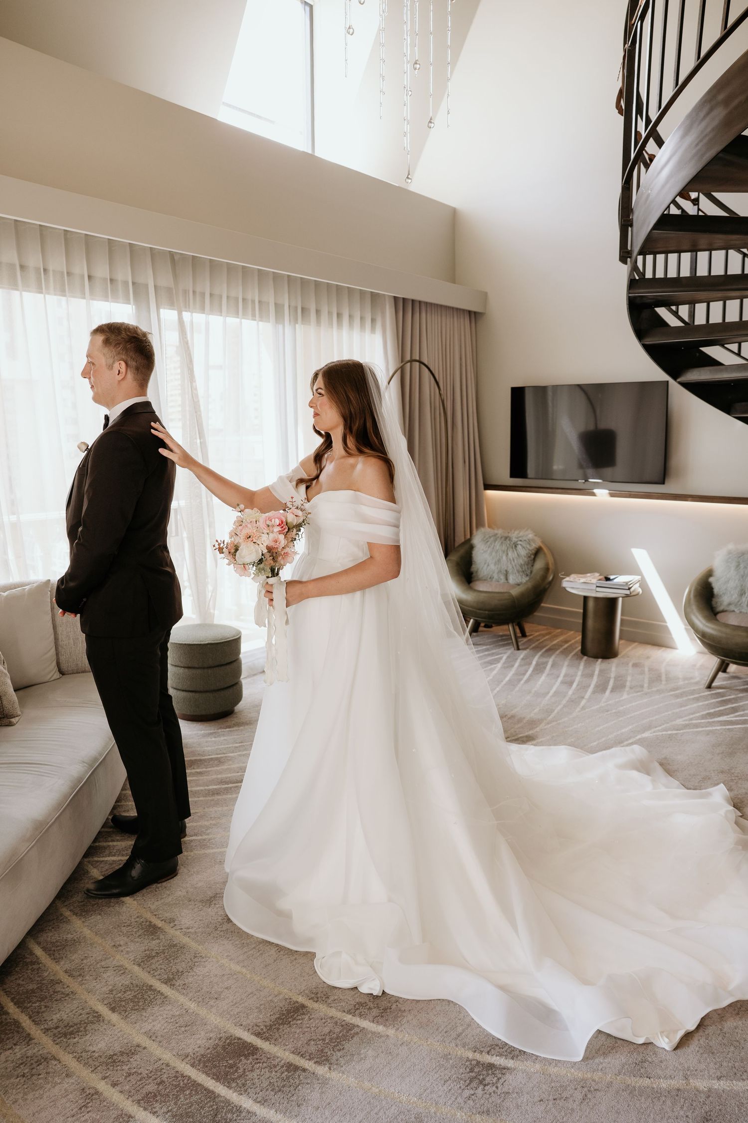 A couple dances together in a bright white room with a spiral staircase, creating a romantic wedding moment.