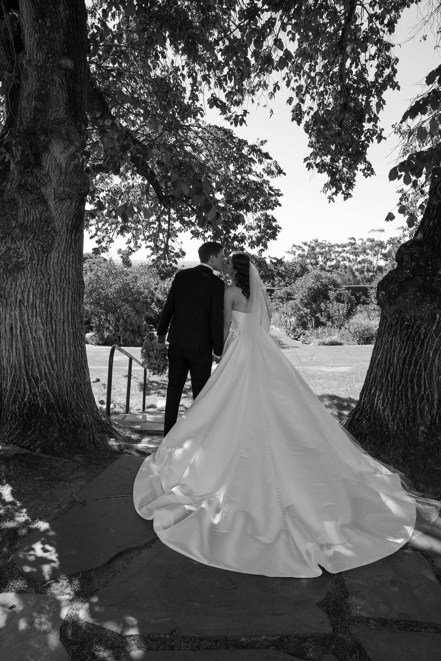 A sequence of black and white wedding photos showing a couple walking together under large trees in a romantic outdoor setting.