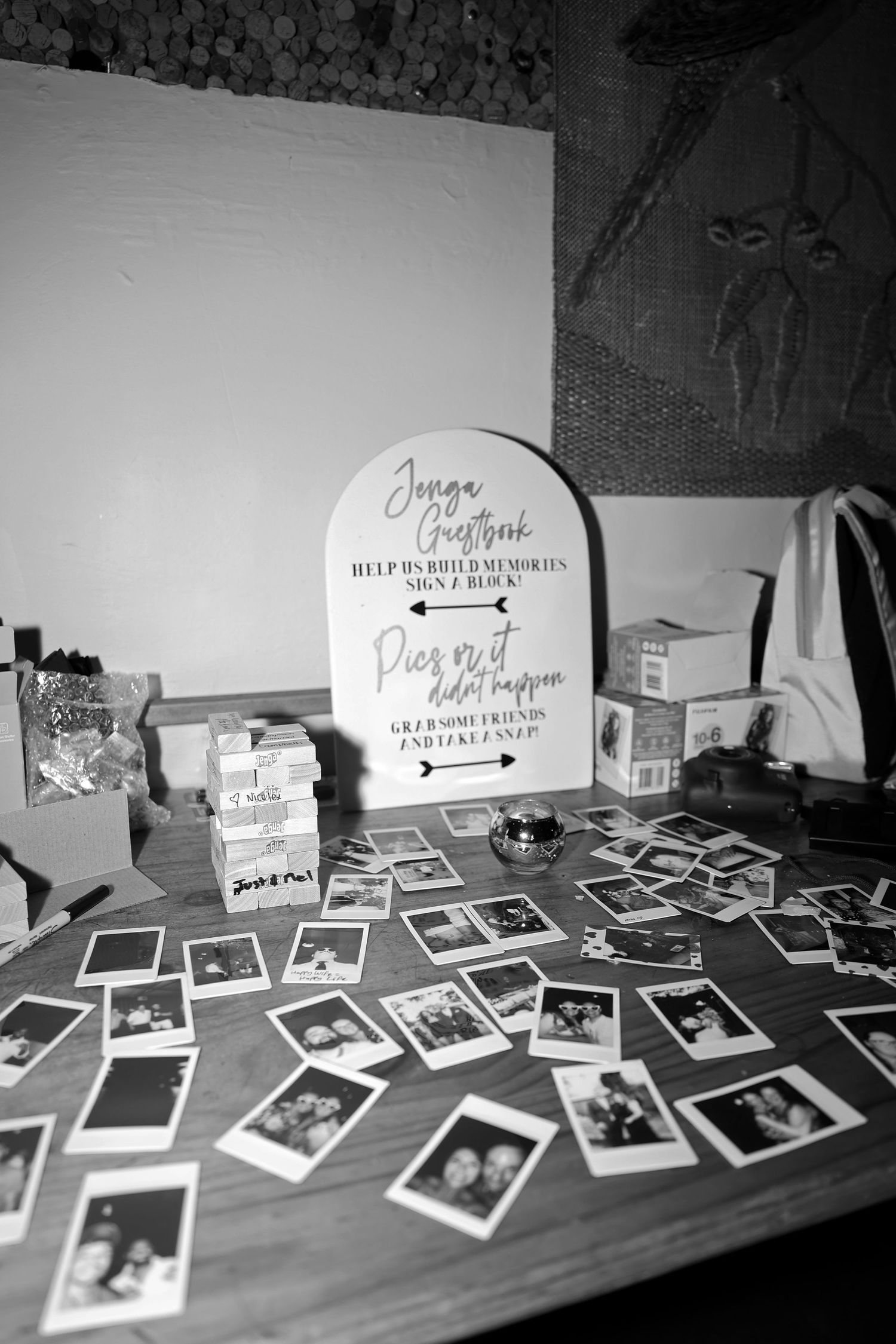 Black and white photo of polaroid photos scattered on a wooden table with a wedding sign display in the background.
