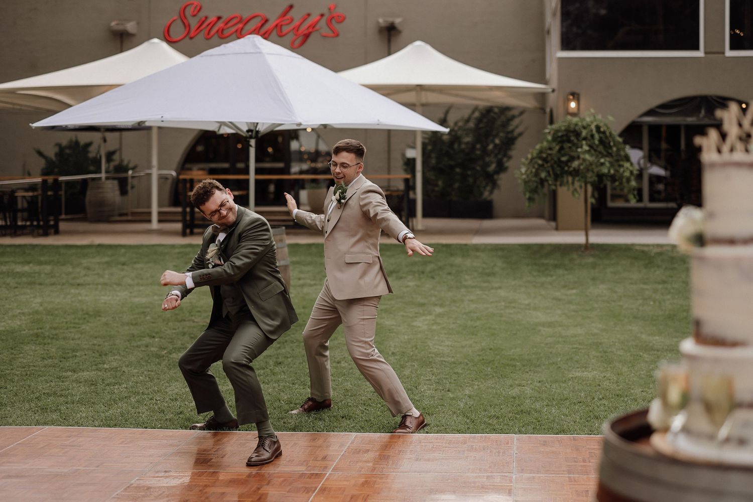 Groomsmen in coordinated suits dance and celebrate under string lights at an outdoor venue.