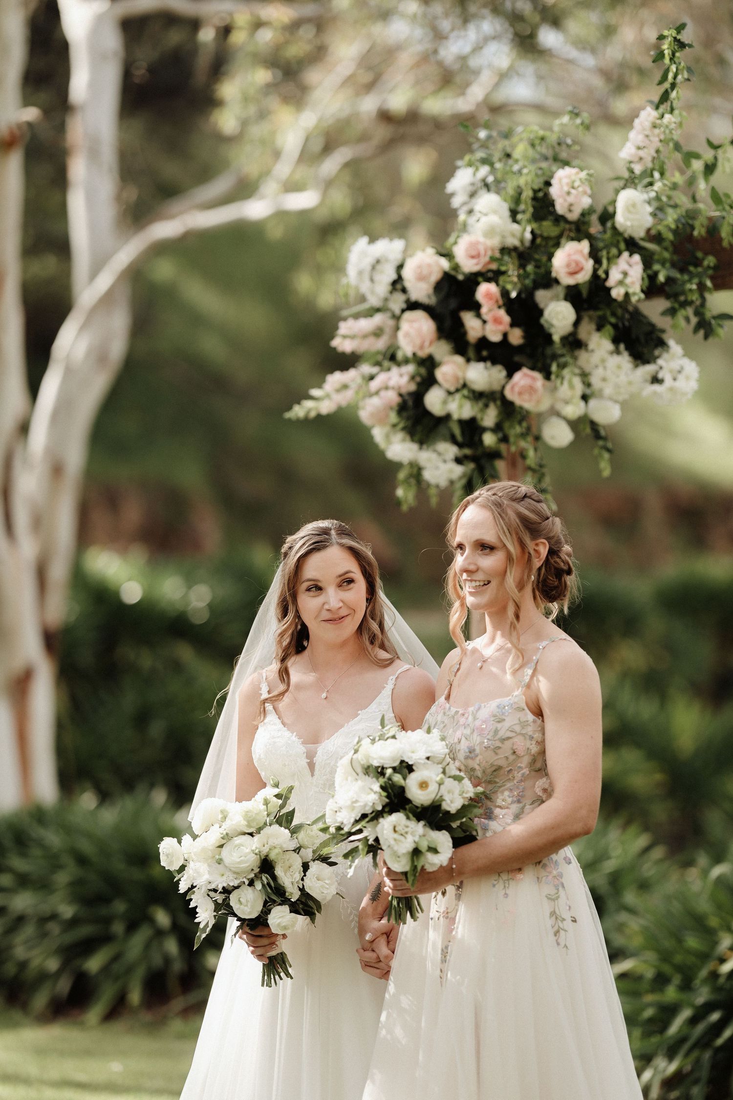 Bridal party in white dresses holds elegant bouquets while posing together in a garden setting.
