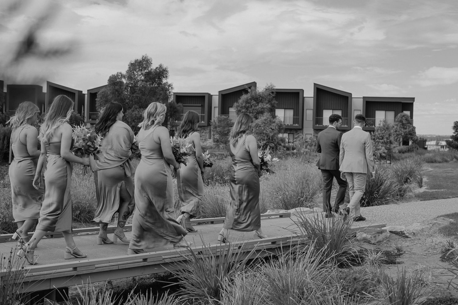 A group of people walk in line across wooden planks in a desert landscape in this vintage black and white photograph.