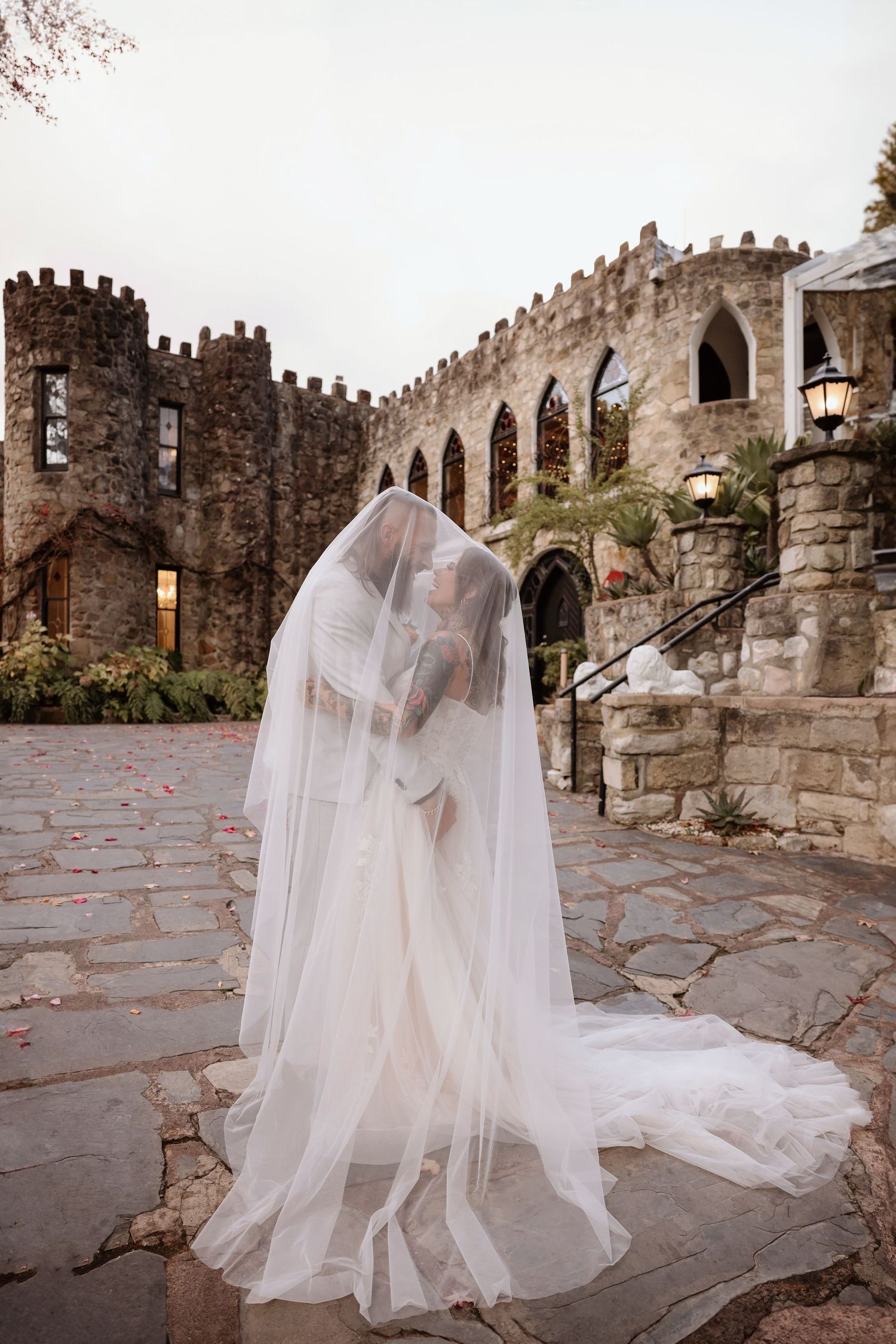 A flowing white wedding dress and veil cascade across stone pavement against medieval castle walls and archways.