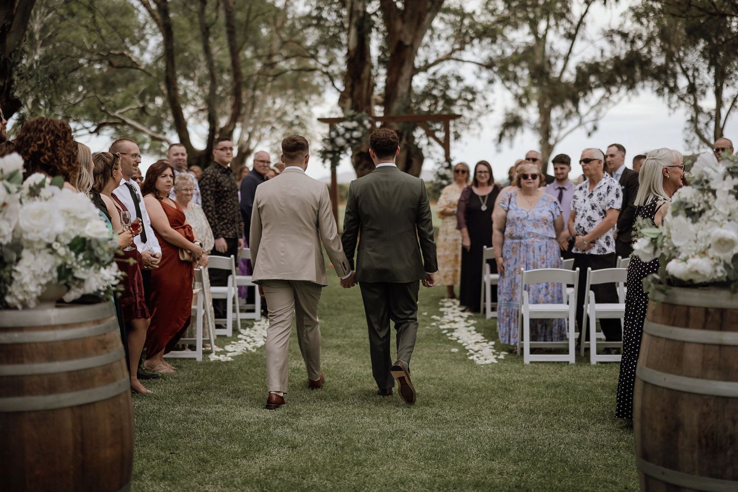 Outdoor wedding ceremony setup with guests gathered on a grassy path lined with rustic wine barrels and white chairs.