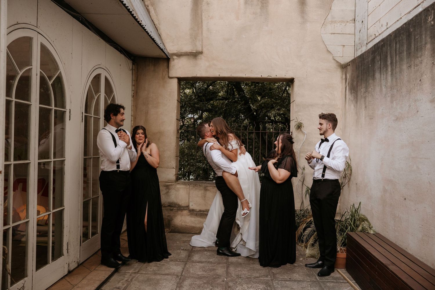 Wedding party stands in formal attire against a light stone wall at an elegant venue.