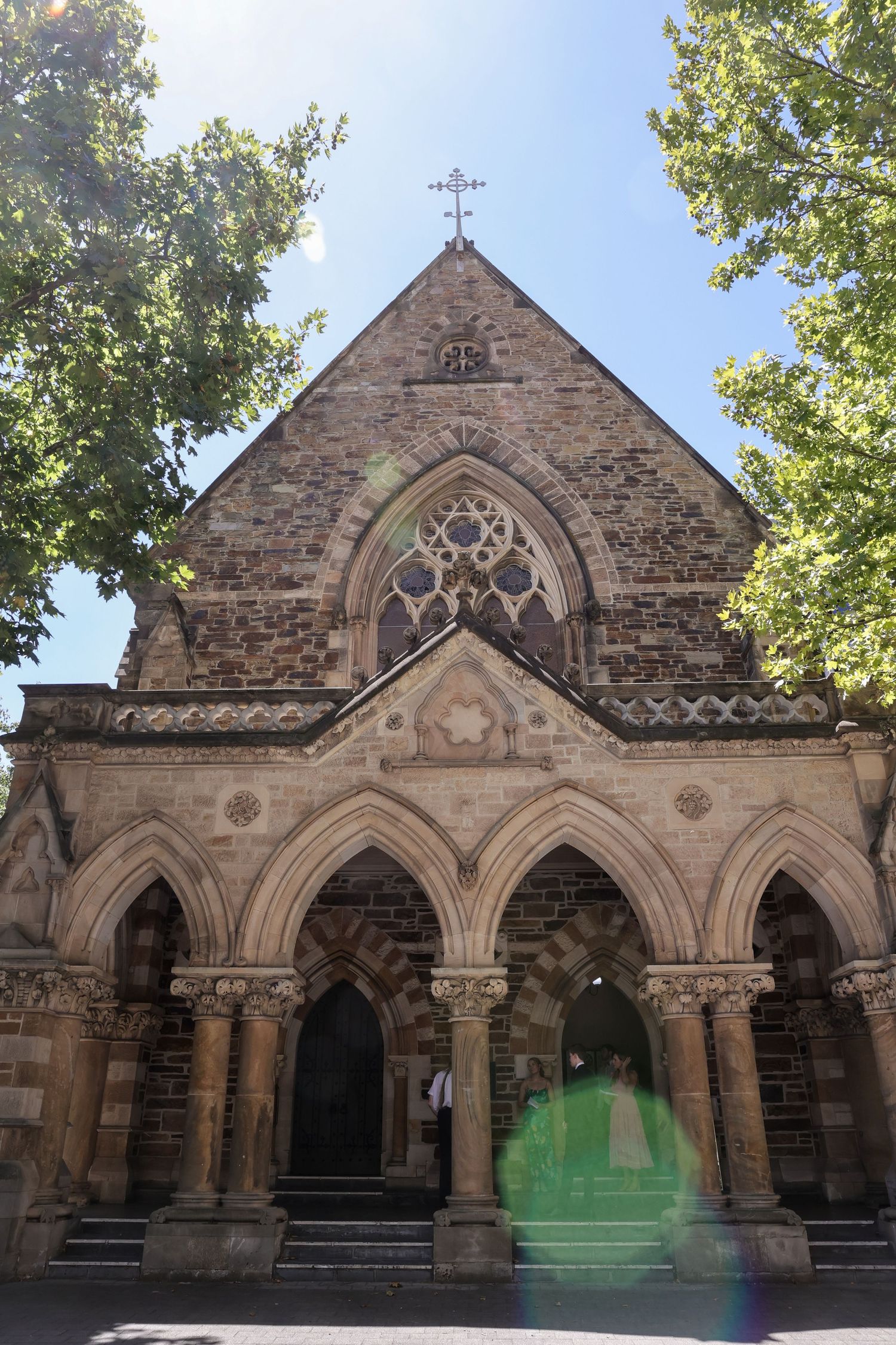 Gothic stone church entrance with ornate arched doorways, columns and cross on top, framed by green trees against blue sky.