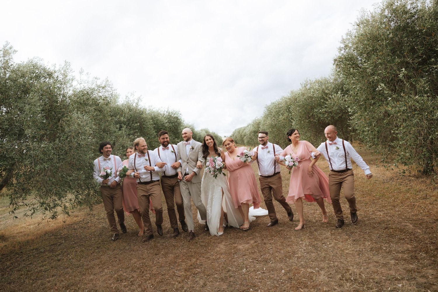 Wedding party in casual attire poses along a rustic dirt path surrounded by olive trees on a sunny day.