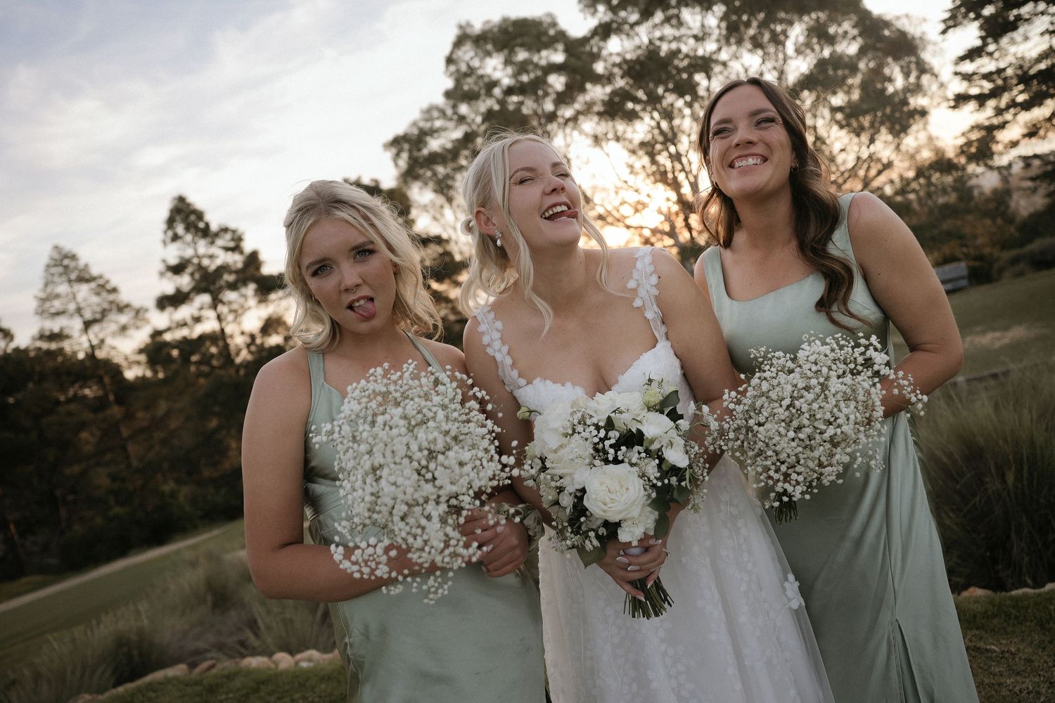 Bridesmaids in sage green dresses share a joyful moment together outdoors at sunset.