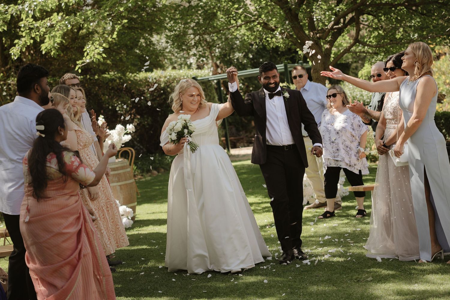 Wedding party celebrates outdoors in a garden setting with guests throwing flower petals during summer festivities.