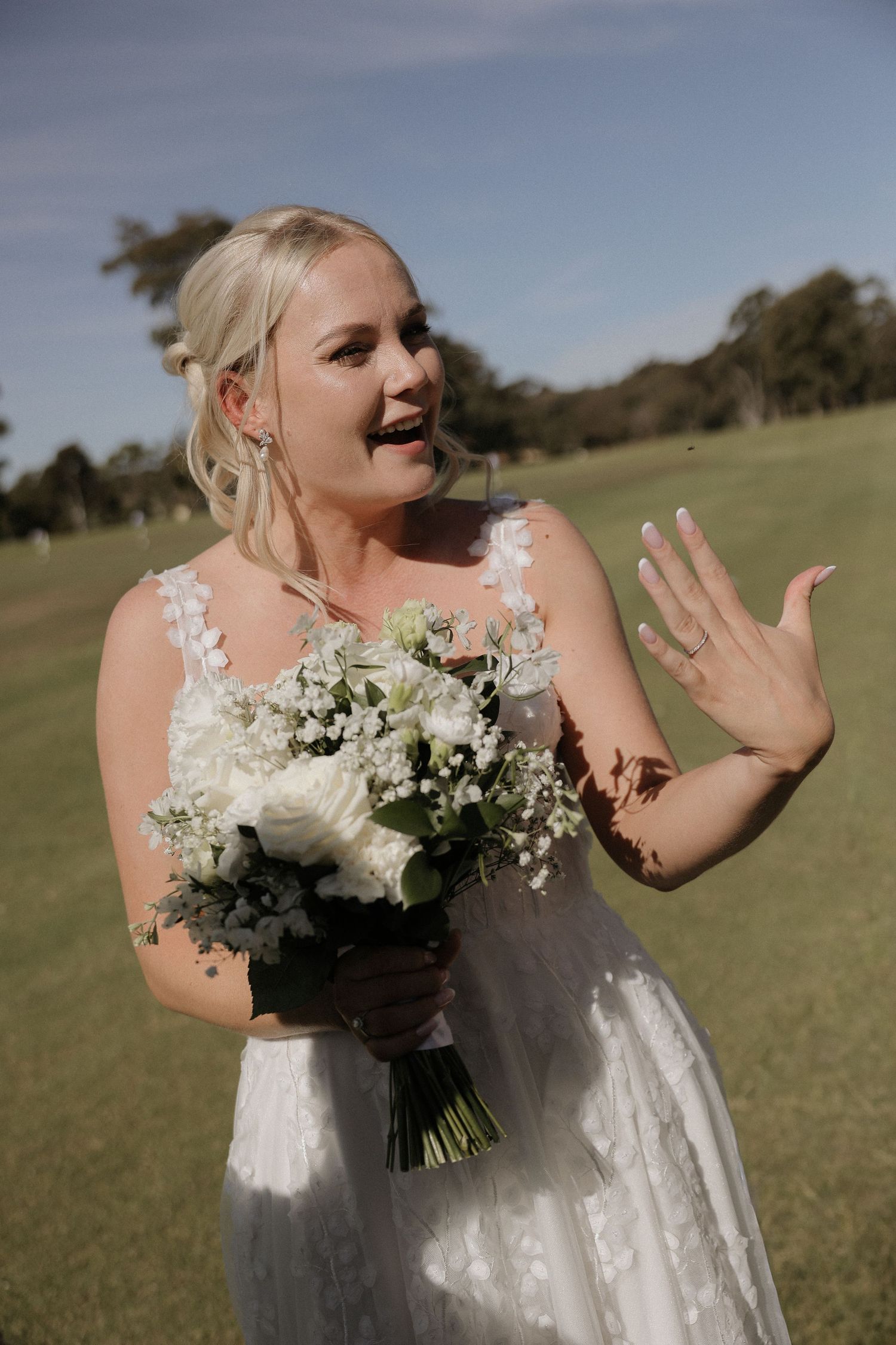 A bride in a white dress holds a bouquet of white flowers while making animated gestures in an outdoor field setting.
