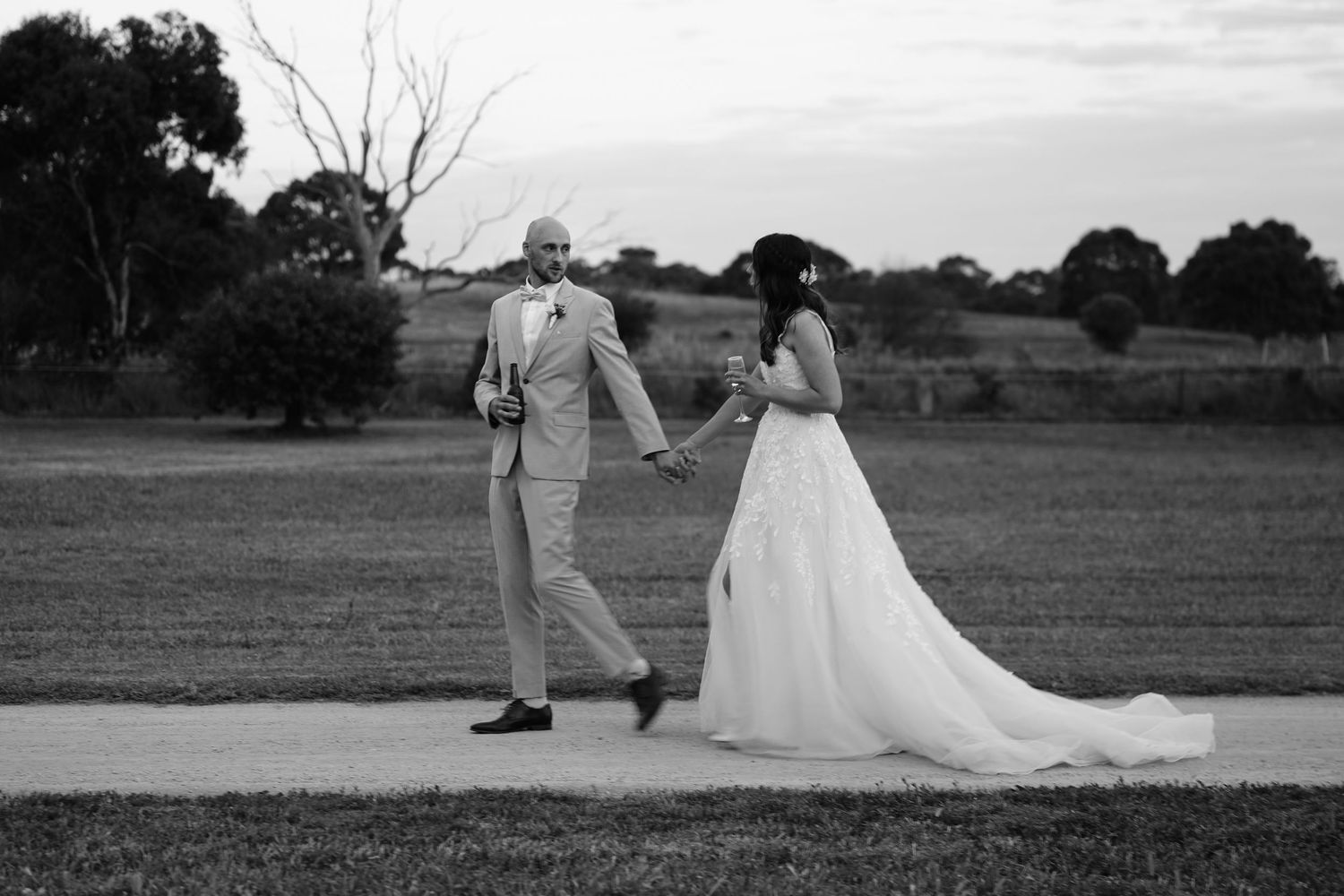 A black and white wedding photograph showing a couple holding hands on a path in an outdoor countryside setting.