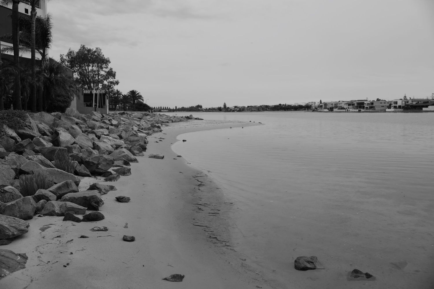 Rocky shoreline curves along a calm ocean bay with trees and buildings visible in the distance.