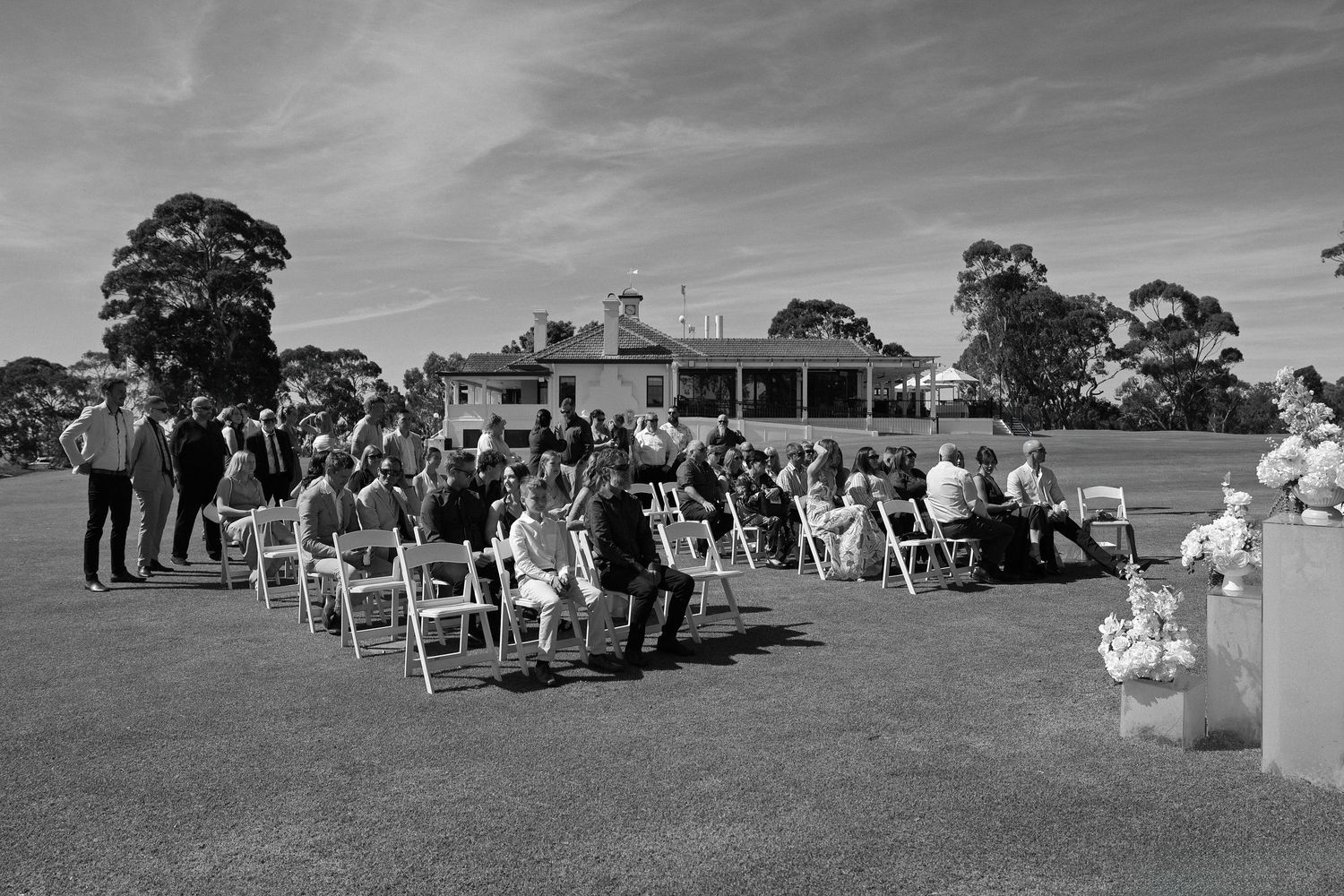 A panoramic black and white photograph shows an outdoor wedding ceremony setup with rows of chairs on a grassy field.
