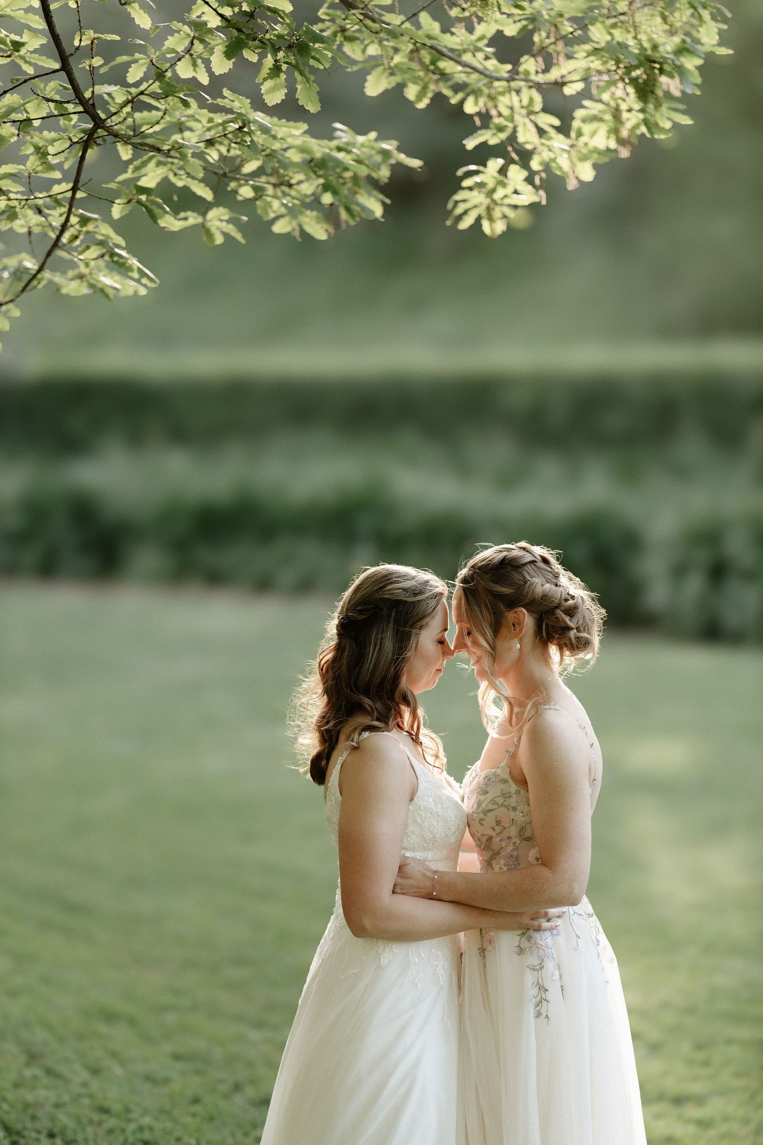 Two brides in white wedding dresses share an intimate moment outdoors under blooming tree branches.