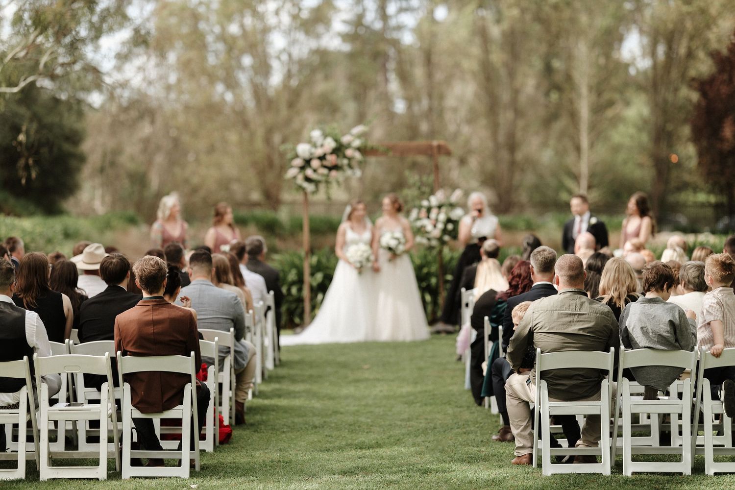 A panoramic view of an outdoor wedding ceremony with white chairs arranged on a lush green lawn surrounded by trees.