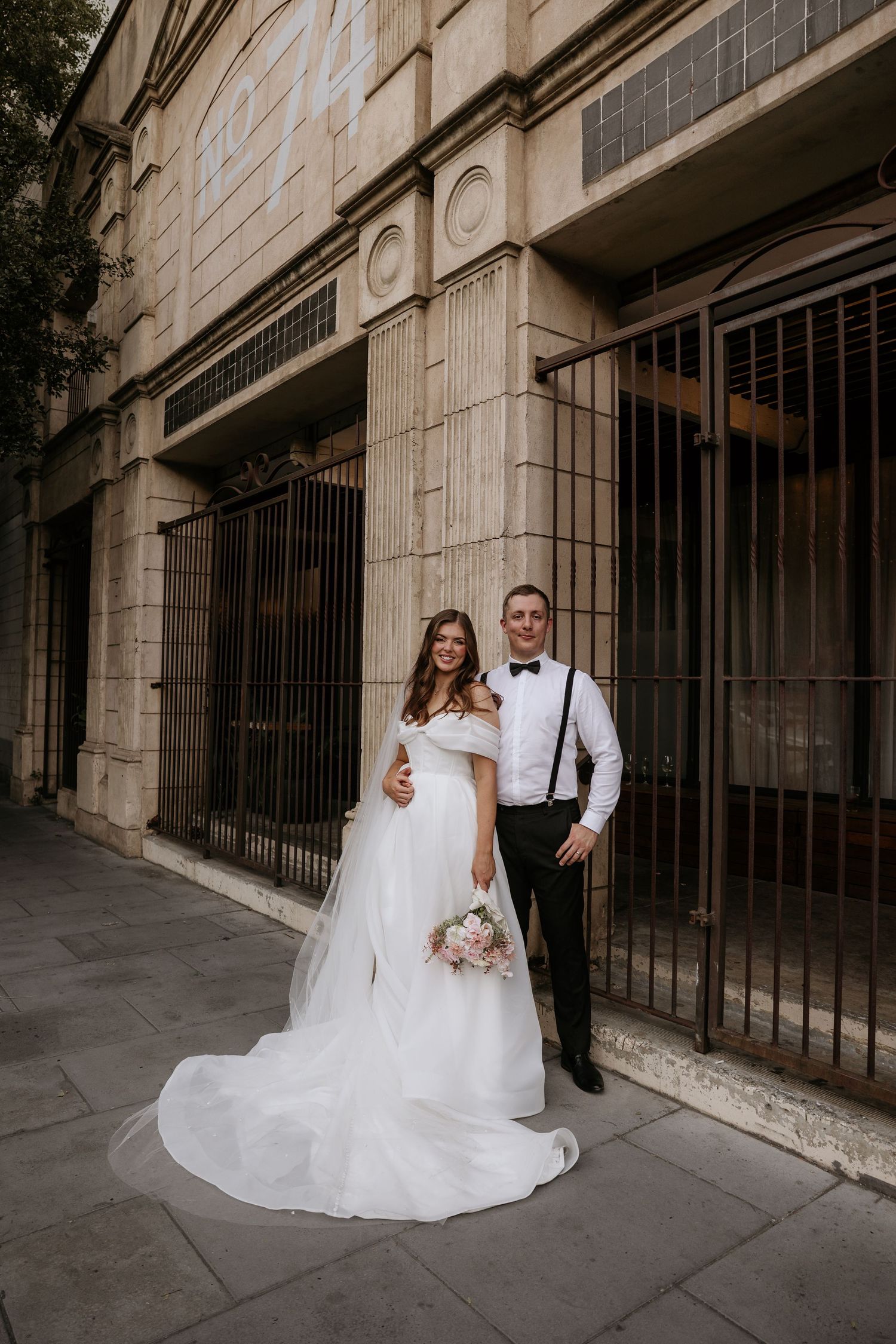 A wedding couple poses together against a stone building wall with black iron bars in an urban setting.
