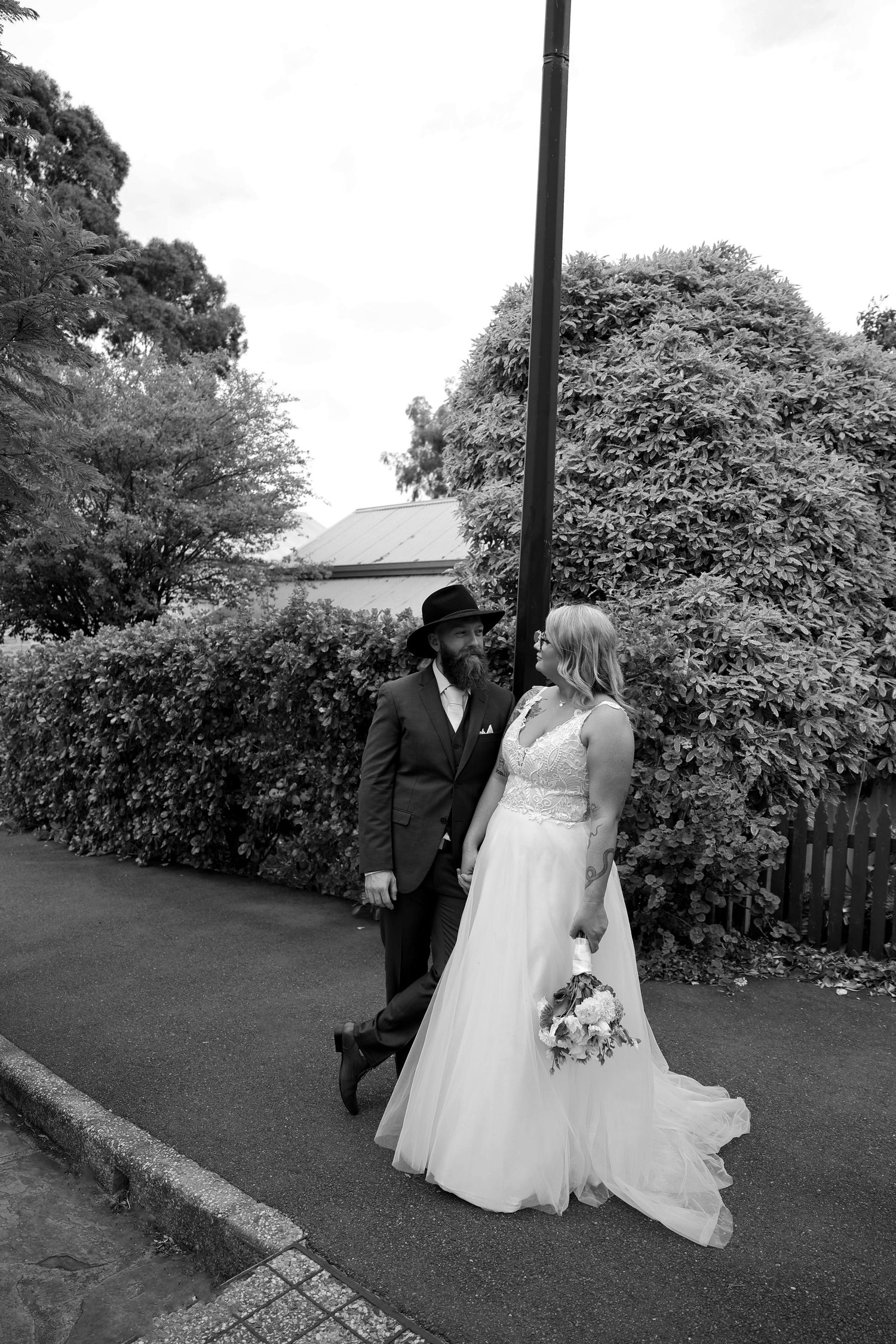 A wedding couple walks along a lush garden path in black and white, with trees and hedges creating a romantic backdrop.