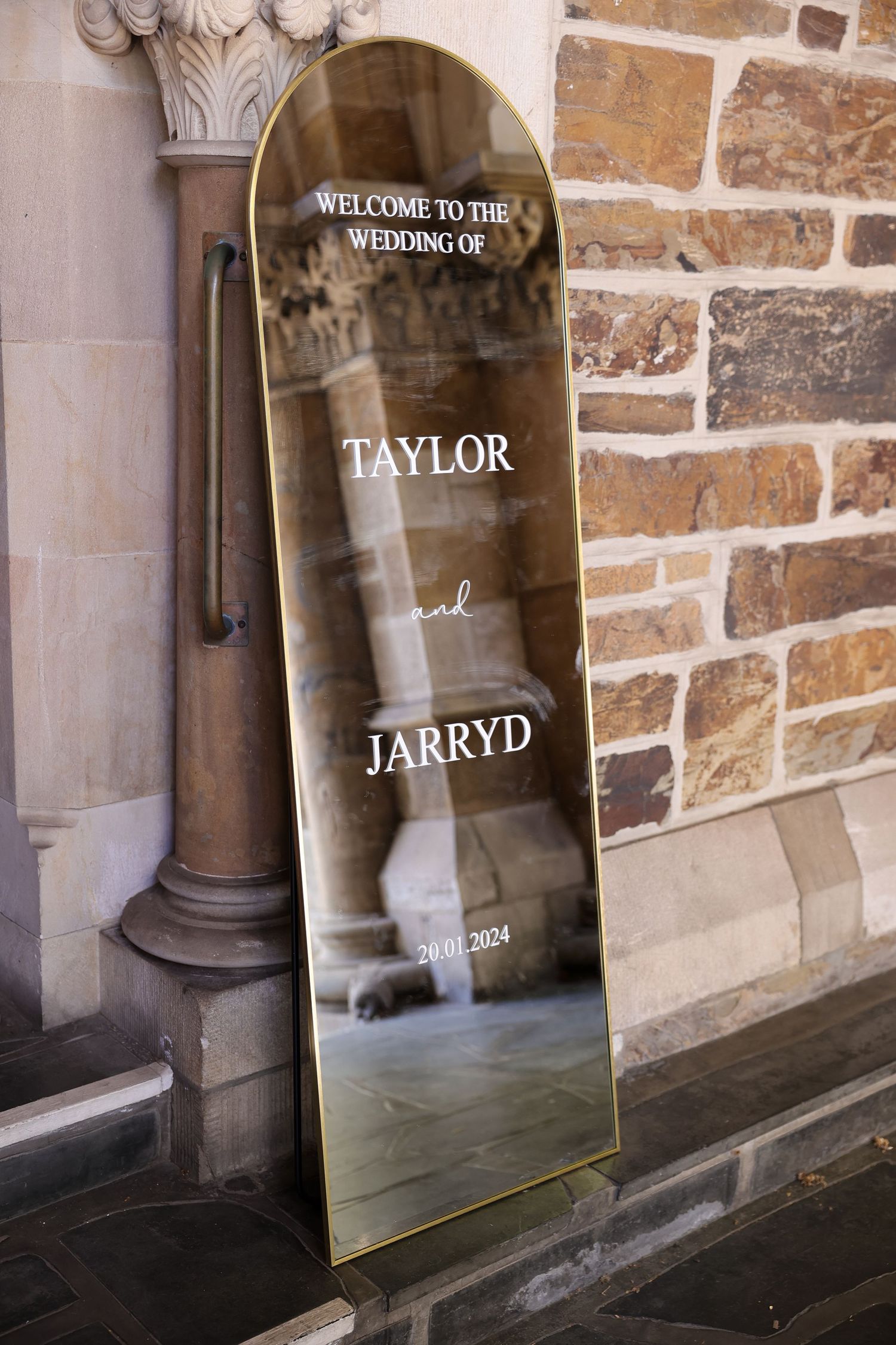Modern acrylic welcome sign for a wedding displayed against a rustic stone wall interior.