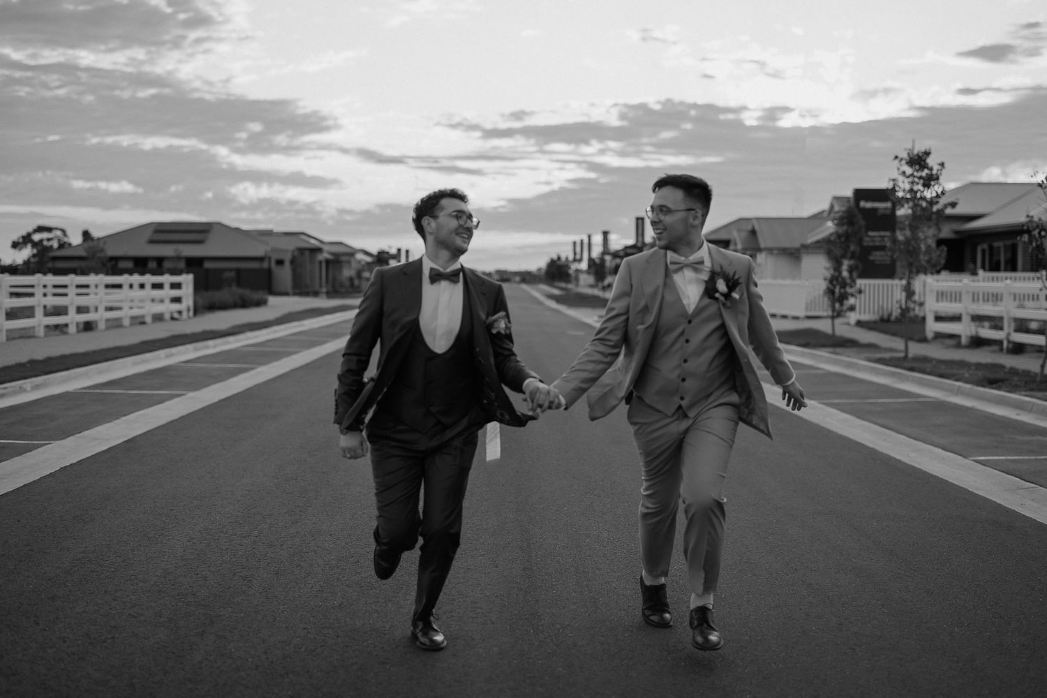 Two people in formal attire run joyfully hand-in-hand down a street with mountains and fences in the background.